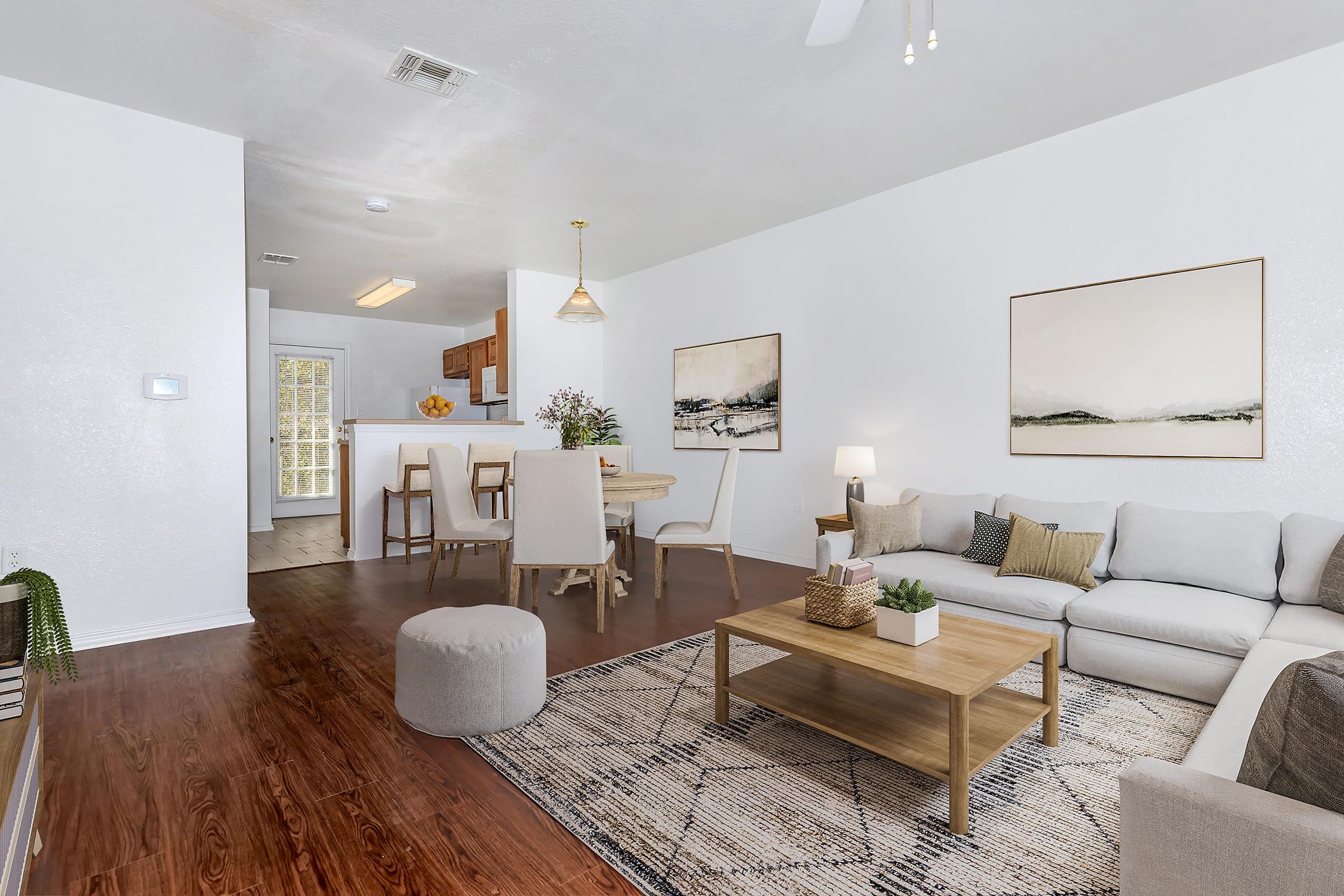 Cozy living room featuring a light gray sectional sofa with decorative pillows, a wooden coffee table, and a plush ottoman. Natural light fills the space, complemented by two artwork pieces on the walls. A dining area with a round table and chairs is visible in the background, set against a neutral color palette.