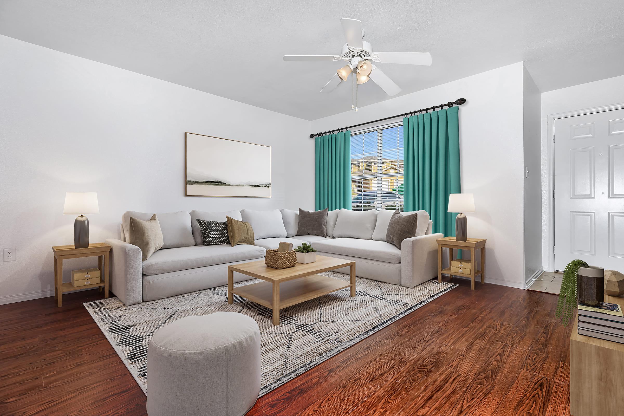 A bright and inviting living room featuring a light-colored sofa with decorative pillows, a wooden coffee table, and a round ottoman. Large windows with teal curtains provide natural light, while a neutral rug and minimal decor create a cozy atmosphere. The space has wood flooring and a welcoming feel.