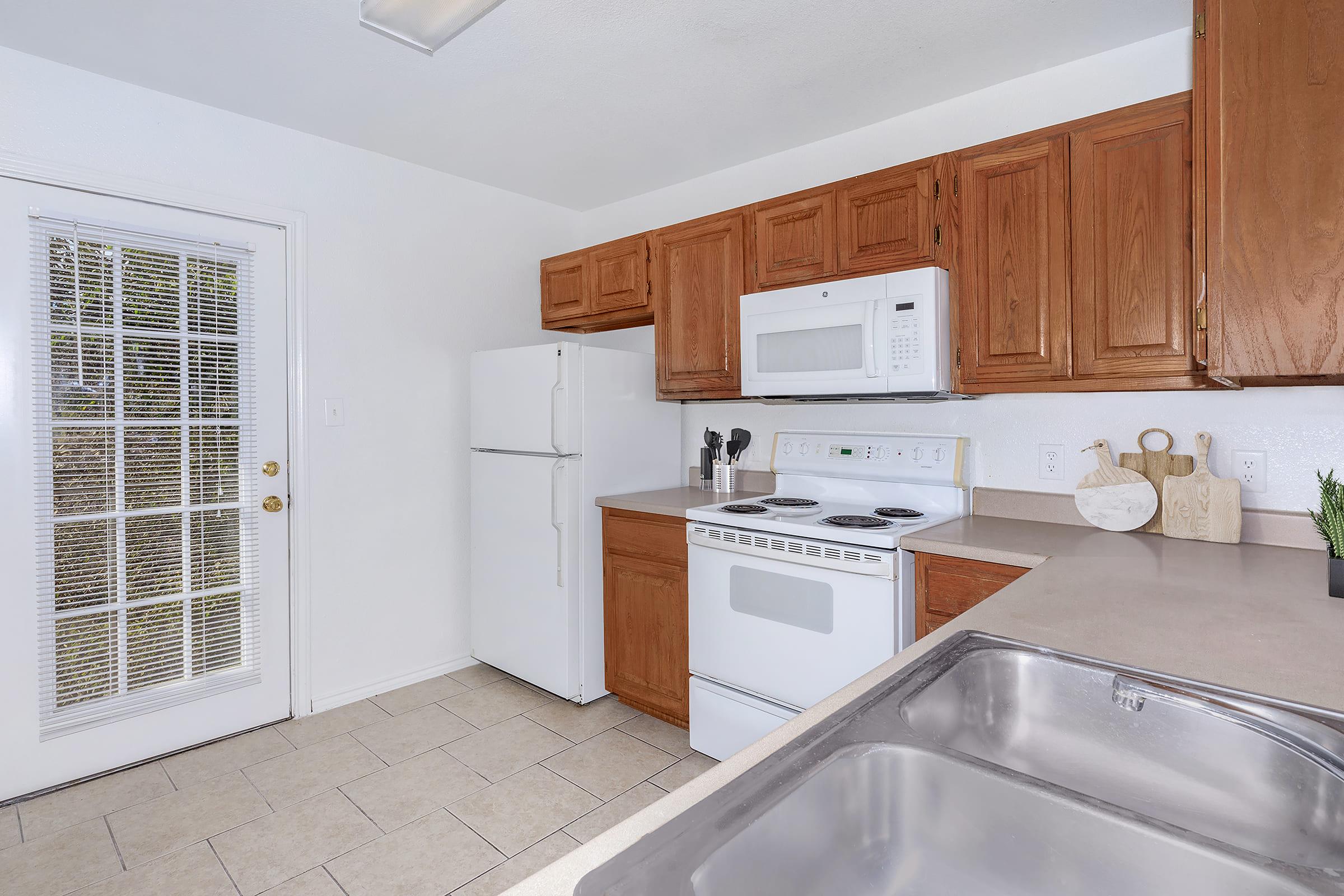 Modern kitchen featuring wooden cabinets, a white refrigerator, a microwave above the stove, and a double basin kitchen sink. Natural light enters through a glass-paned door leading outside. Countertops are clean and uncluttered, enhancing the spaciousness of the room.