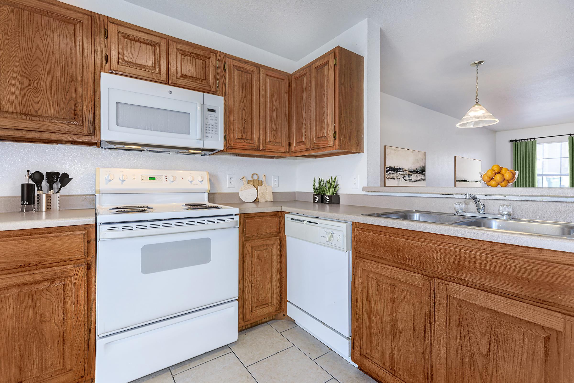 Modern kitchen featuring wooden cabinets, a white stove, and a microwave above. There's a dishwasher on the right and a double sink with a modern faucet. Natural light pours in from a nearby window, with a light fixture hanging above. A bowl of oranges sits on the counter, adding a pop of color to the space.