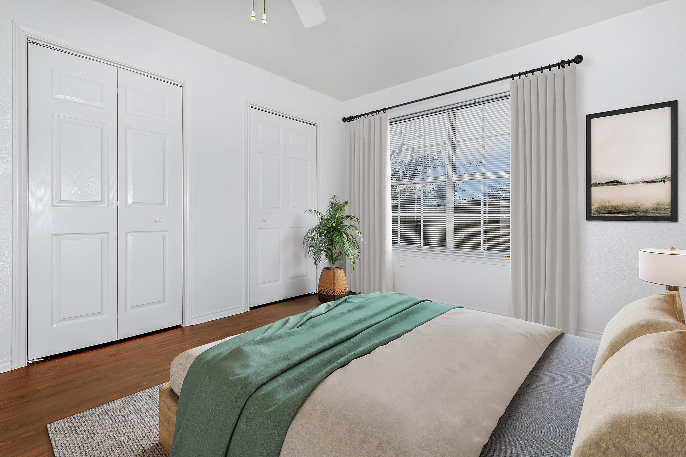 A cozy bedroom featuring a neatly made bed with a green blanket. There are two closet doors on one wall, a large window with sheer curtains allowing natural light, a potted plant in a decorative basket, and a framed artwork hanging on the wall, adding a touch of elegance to the space.