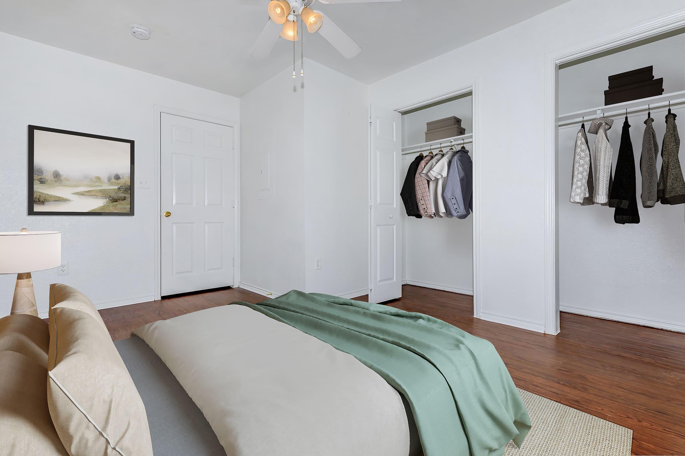 A well-lit bedroom featuring a neatly made bed with a light green blanket, a bedside lamp, and a wall art piece. There are two closets with hanging clothes on one side and a white door leading out. The wooden floor adds warmth to the space, creating a cozy atmosphere.