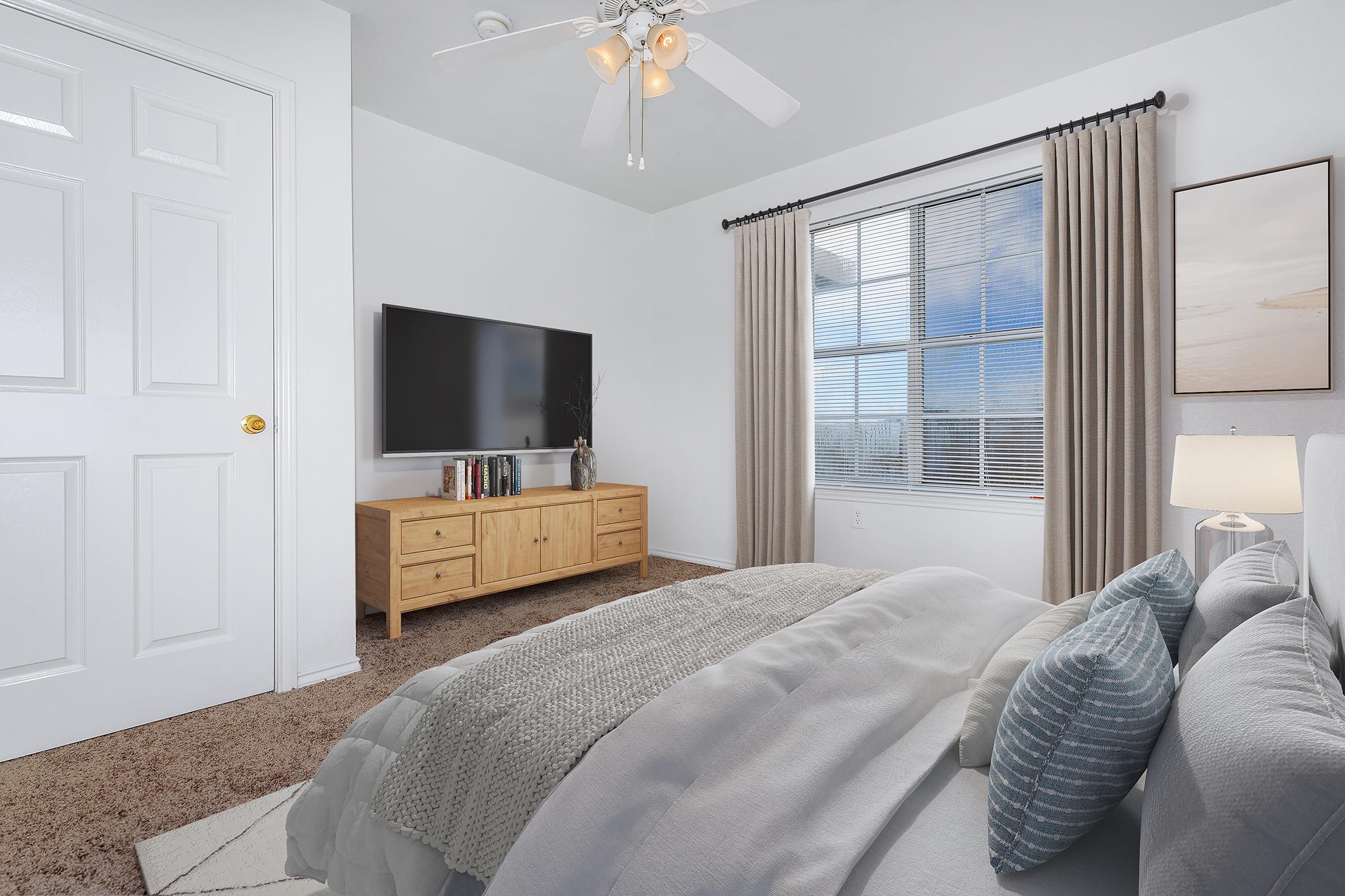 A cozy bedroom featuring a large window with blinds, light-colored walls, and a ceiling fan. A neatly made bed is in the foreground, accompanied by decorative pillows. A modern wooden dresser holds books and a TV screen, while soft curtains frame the window, creating a warm and inviting atmosphere.