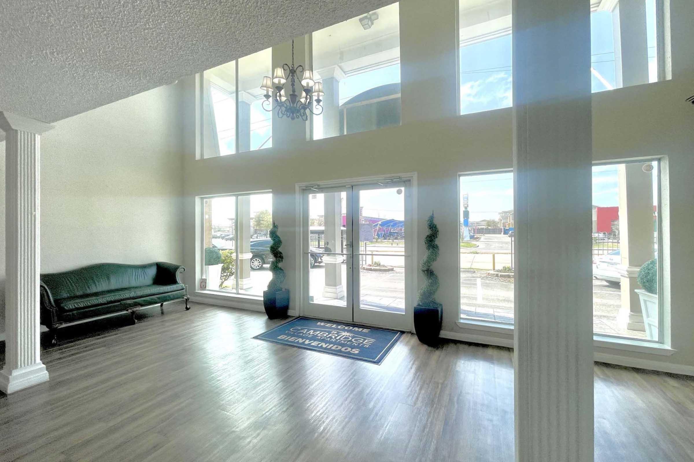 Interior of a bright, spacious lobby featuring large windows allowing natural light. A dark green sofa sits against a wall, and two decorative planters stand near the entrance. The floor is light-colored wood, and a welcome mat is positioned at the door.