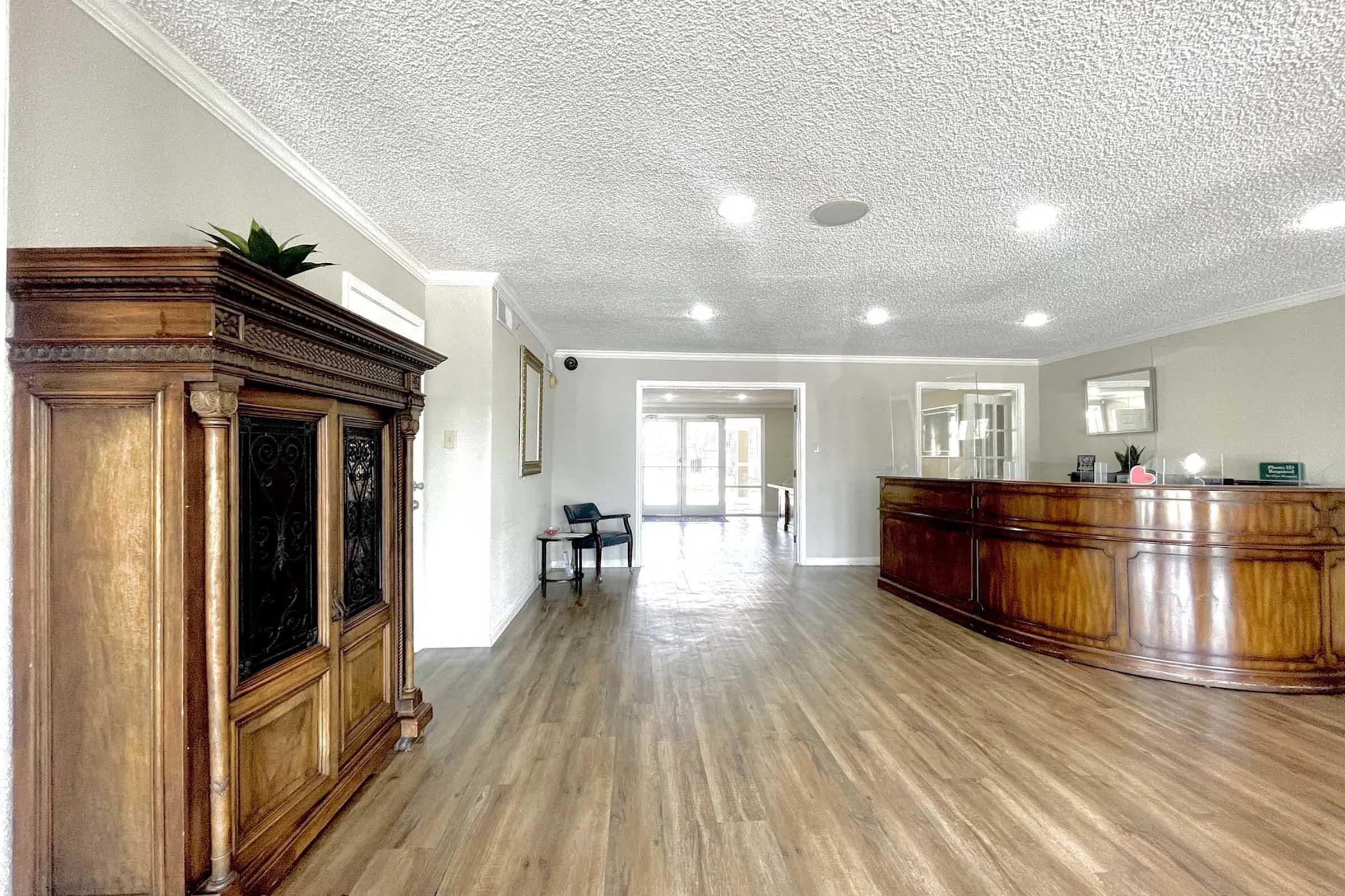 A spacious lobby area featuring a wooden reception desk, a stylish cabinet on the left, and light-colored walls. The floor is covered with hardwood, and bright ceiling lights illuminate the space. Large windows at the far end allow natural light to enter, creating a welcoming atmosphere.