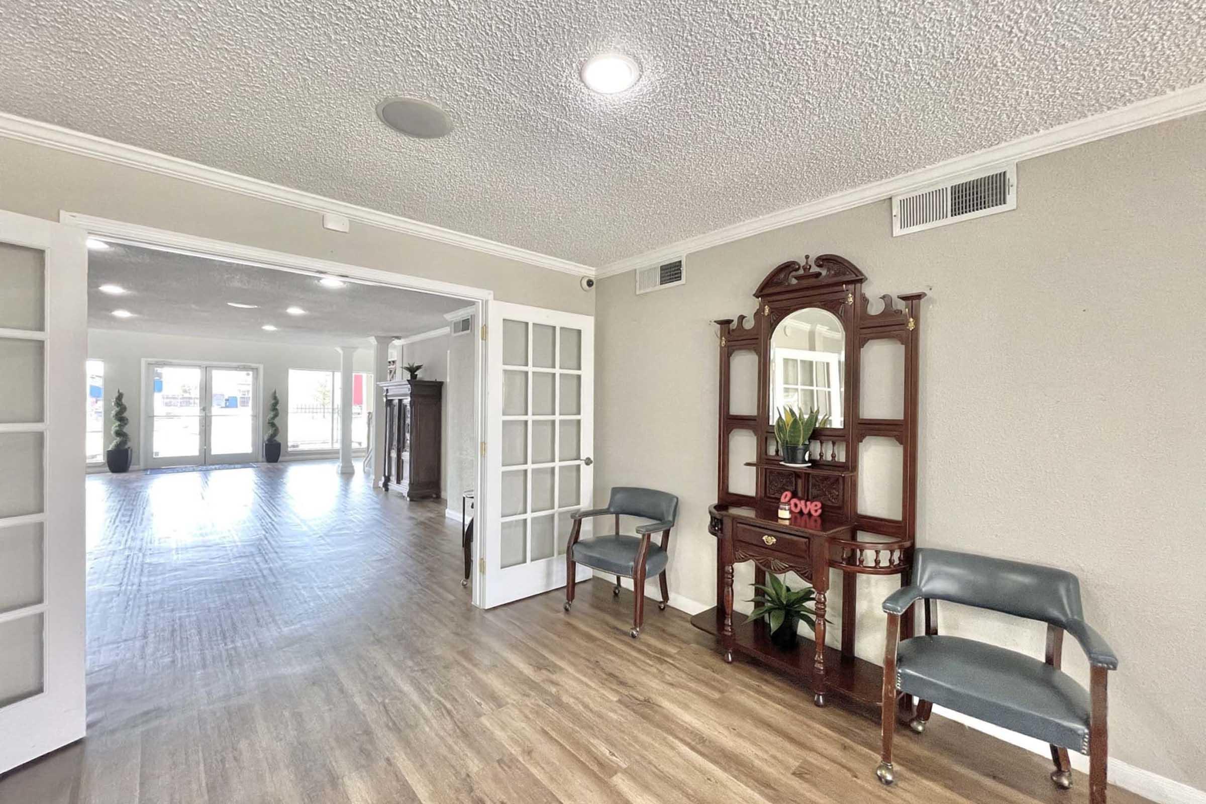 A welcoming interior space featuring a hardwood floor and neutral-colored walls. On the left, there are two blue chairs and a vintage wooden console table with a small plant. The area is well-lit, creating a warm atmosphere, and there are glass-paned doors leading to a larger room in the background.