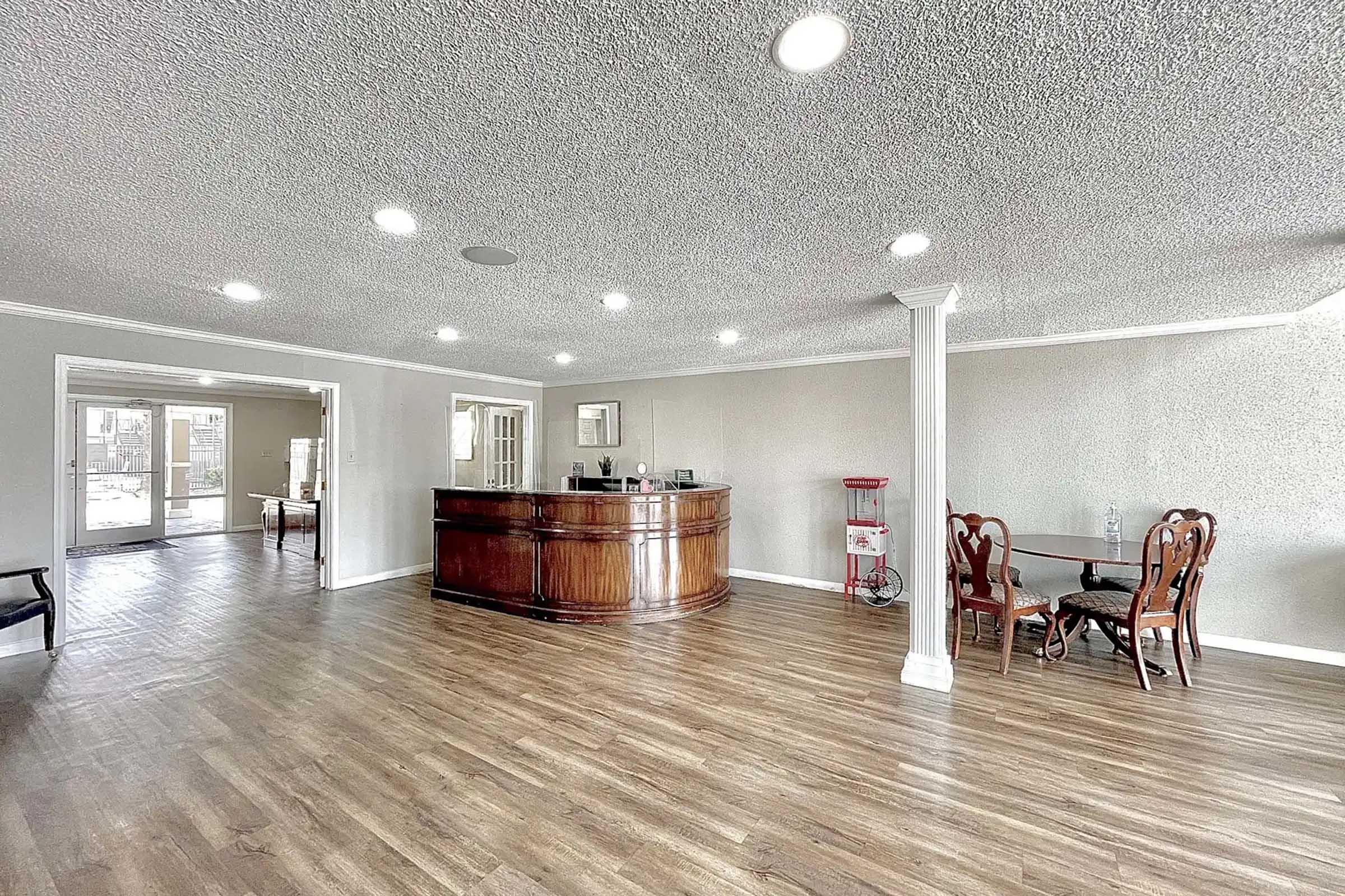 Interior view of a spacious lobby area featuring a wooden reception desk, a small round dining table with four chairs, and large windows allowing natural light. The flooring is wood-like, and the ceiling has recessed lighting, creating a welcoming atmosphere.