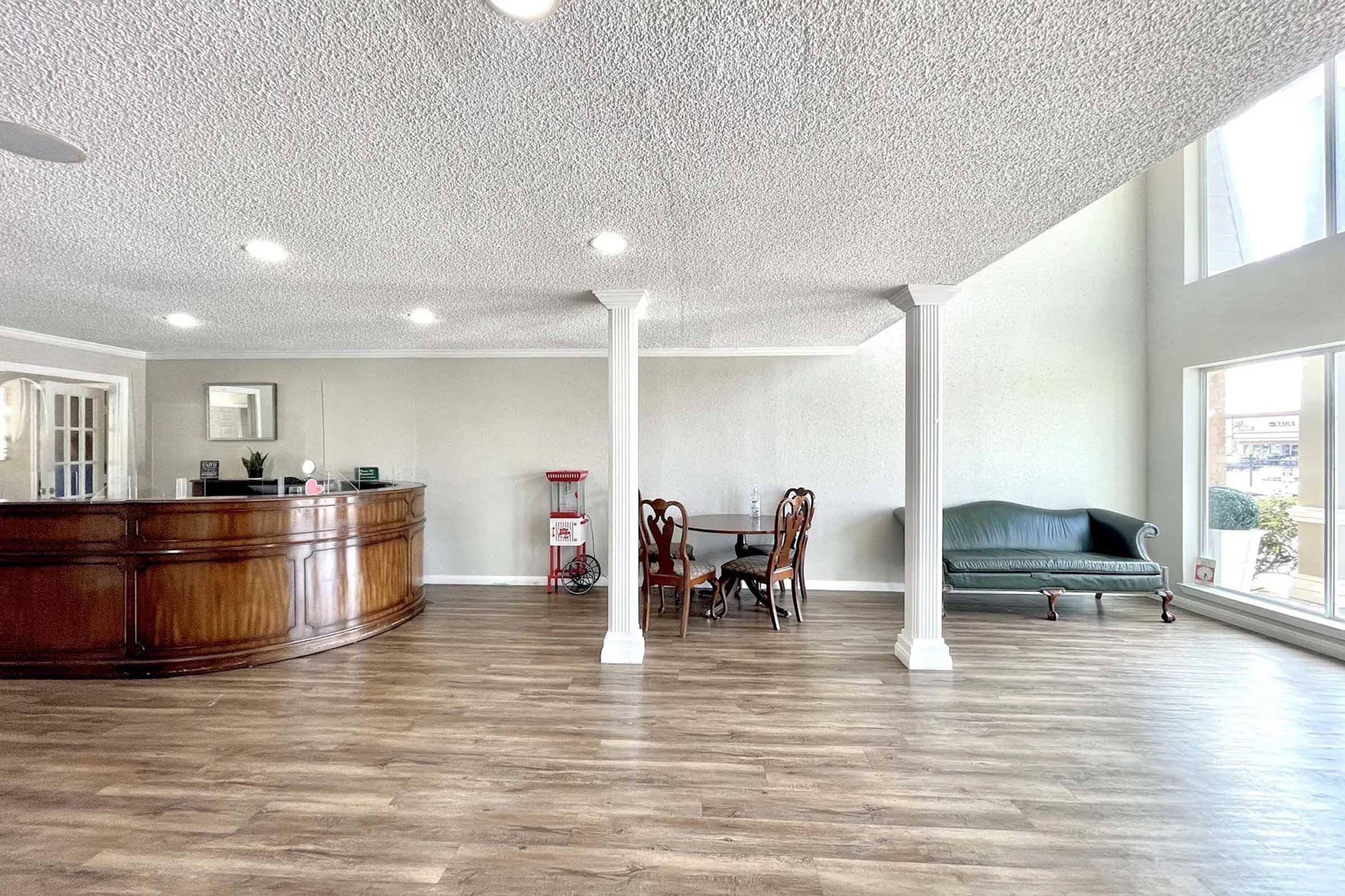 A spacious reception area featuring a wooden front desk, a small dining table with chairs, and a green couch. Brightly lit with natural light coming through large windows, the space has a modern design and light-colored walls, creating a welcoming atmosphere.