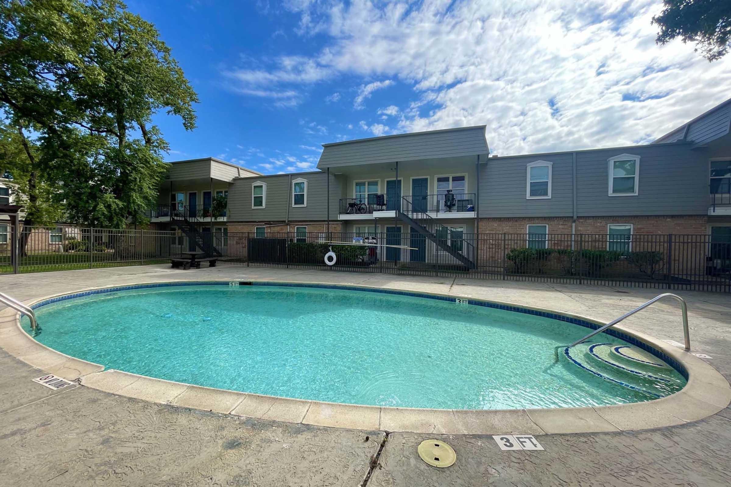 A clear swimming pool surrounded by a textured concrete deck, with a green tree in the background. Two apartment buildings with balconies are visible behind the pool, and a lifebuoy hangs on the fence. The sky is partly cloudy with blue patches.