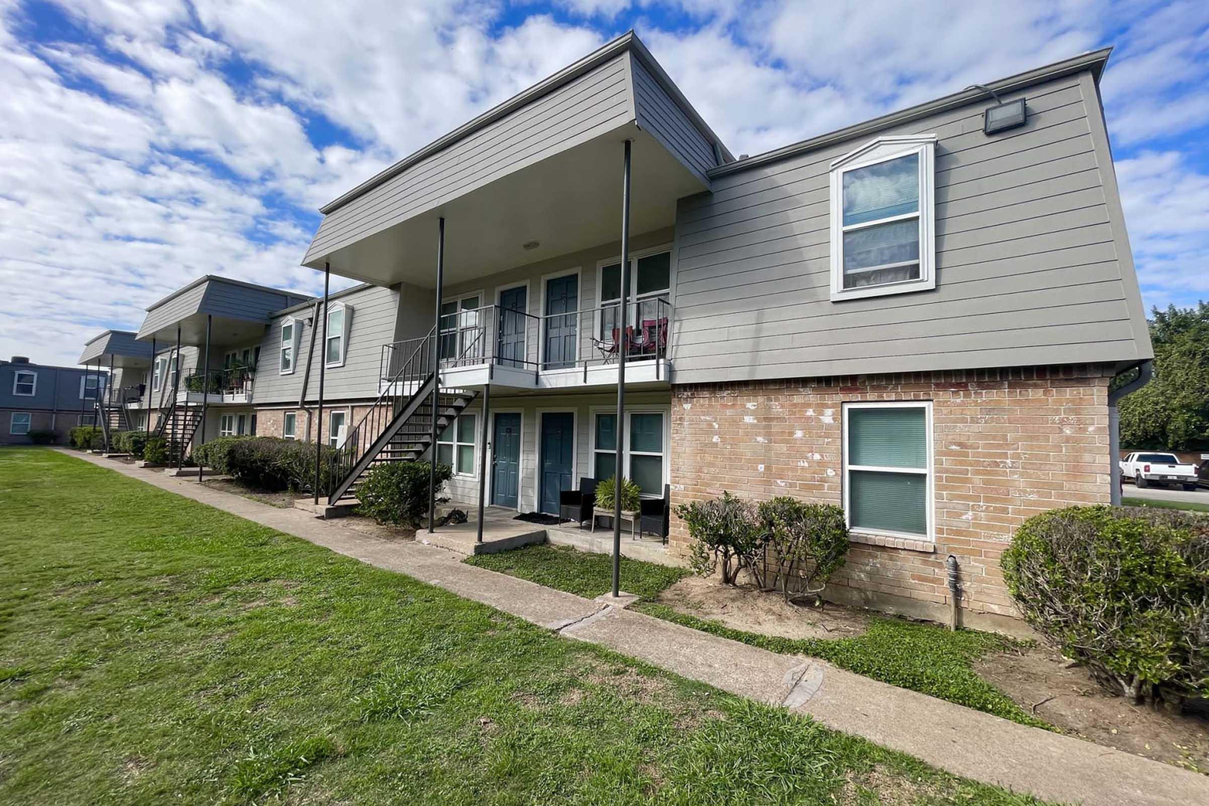 A view of a two-story apartment building with multiple units. Each unit features a balcony and outdoor staircases. The building has a brick base with gray siding and is surrounded by well-maintained grassy areas and shrubs. The sky is partly cloudy, suggesting a clear day.