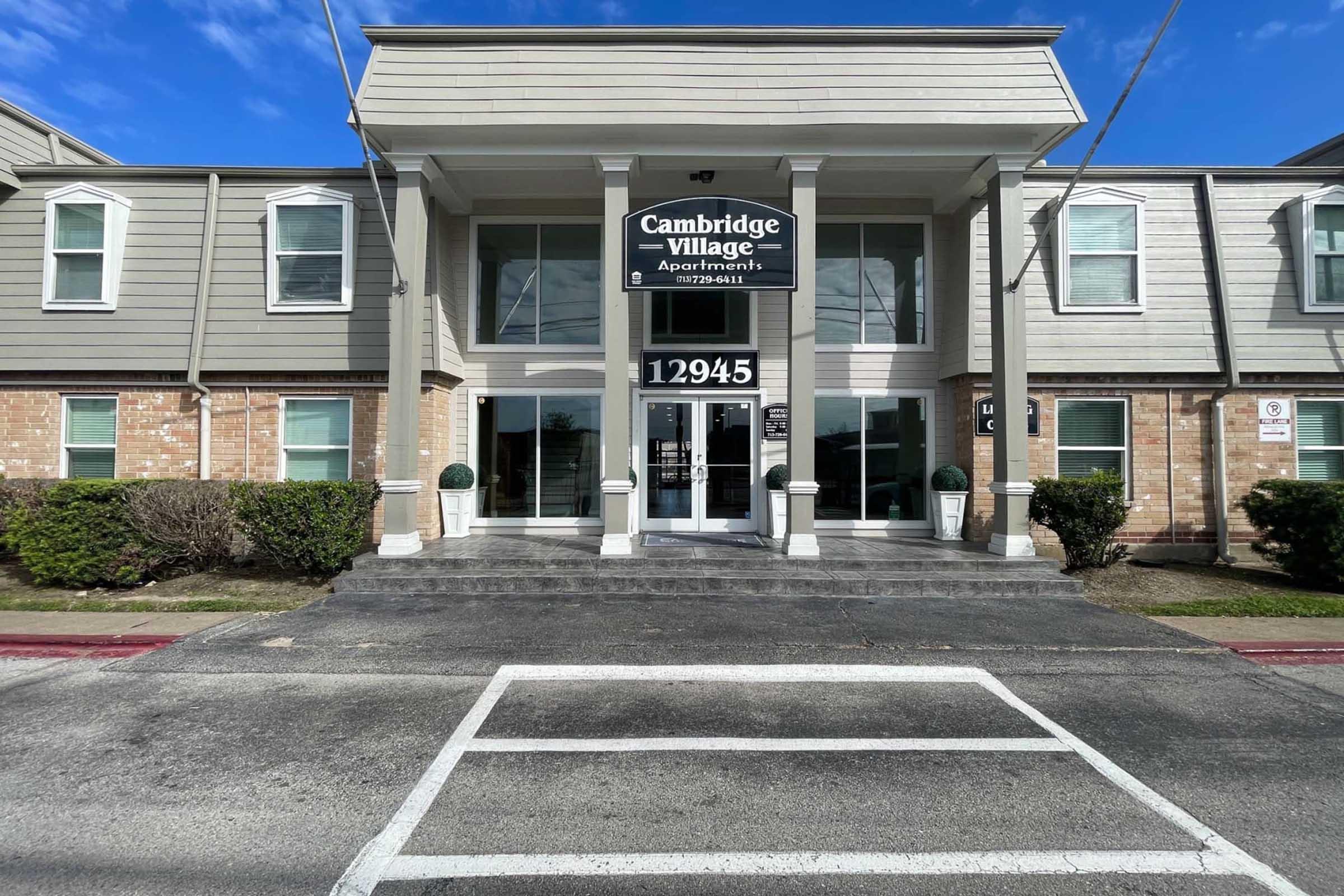 Front entrance of Cambridge Village Apartments, featuring a welcoming facade with large glass doors, signage displaying the apartment name and contact information, and neatly trimmed bushes on either side of the entrance. The building is two stories with a light-colored exterior and a clear blue sky above.