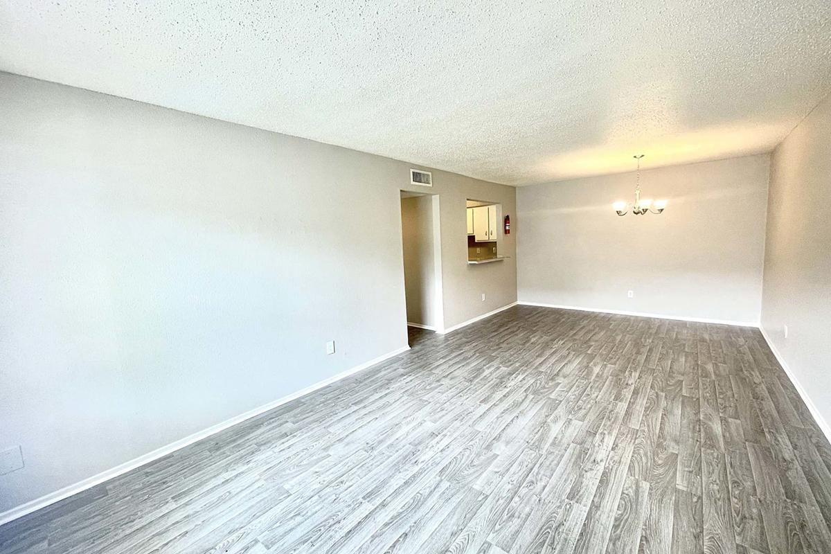 A spacious, empty living room featuring light-colored walls and a wood-like floor. A doorway leads to a kitchen area in the background, and a chandelier hangs above a central dining space. Natural light brightens the room, creating a welcoming atmosphere.