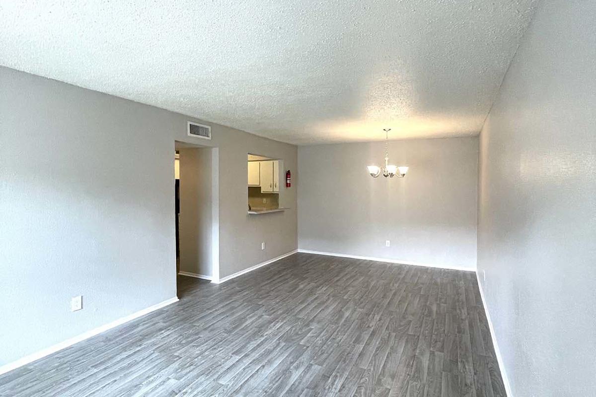 A spacious living room with light gray walls and wood-style laminate flooring. There’s a chandelier hanging from the ceiling, and an adjacent area leads to a small kitchen. The room appears bright and empty, suggesting a recent renovation or preparation for new occupants.