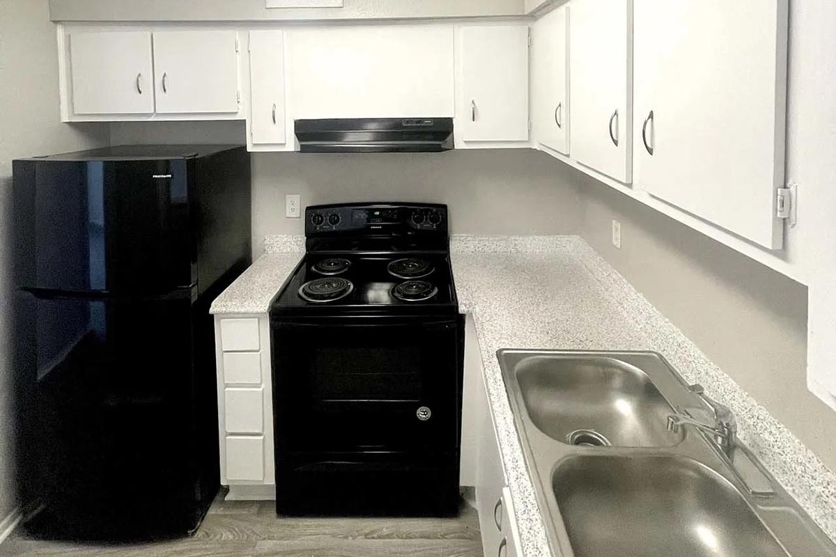 A modern kitchen featuring white cabinets, a black refrigerator, and a black oven with a stovetop. The countertops are light-colored with a speckled design. There are two stainless steel sinks and a window above the sink, providing natural light. The flooring is a light wood tone, creating a contemporary look.