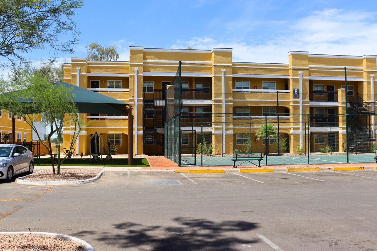 A yellow apartment building with multiple balconies and windows, surrounded by a parking lot. In front, there is a basketball court and a shaded seating area. The sky is blue with a few clouds, and some greenery is visible around the property.