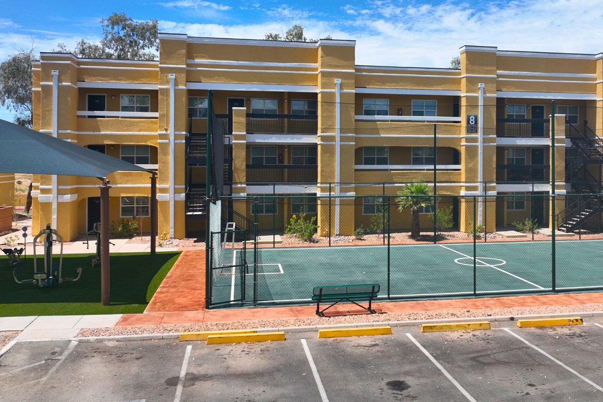 A vibrant yellow apartment building with multiple floors and balconies is visible in the background. In the foreground, there is a basketball court enclosed by a fence, alongside a grassy area and a children's swing set. Clear blue skies are overhead, creating a bright and inviting atmosphere.