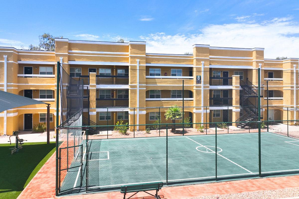 A view of a multi-story yellow residential building with balconies, surrounded by landscaped areas. In the foreground, there's a fenced outdoor basketball court with a green surface and a single basketball hoop. The sky is clear and blue, indicating a sunny day.