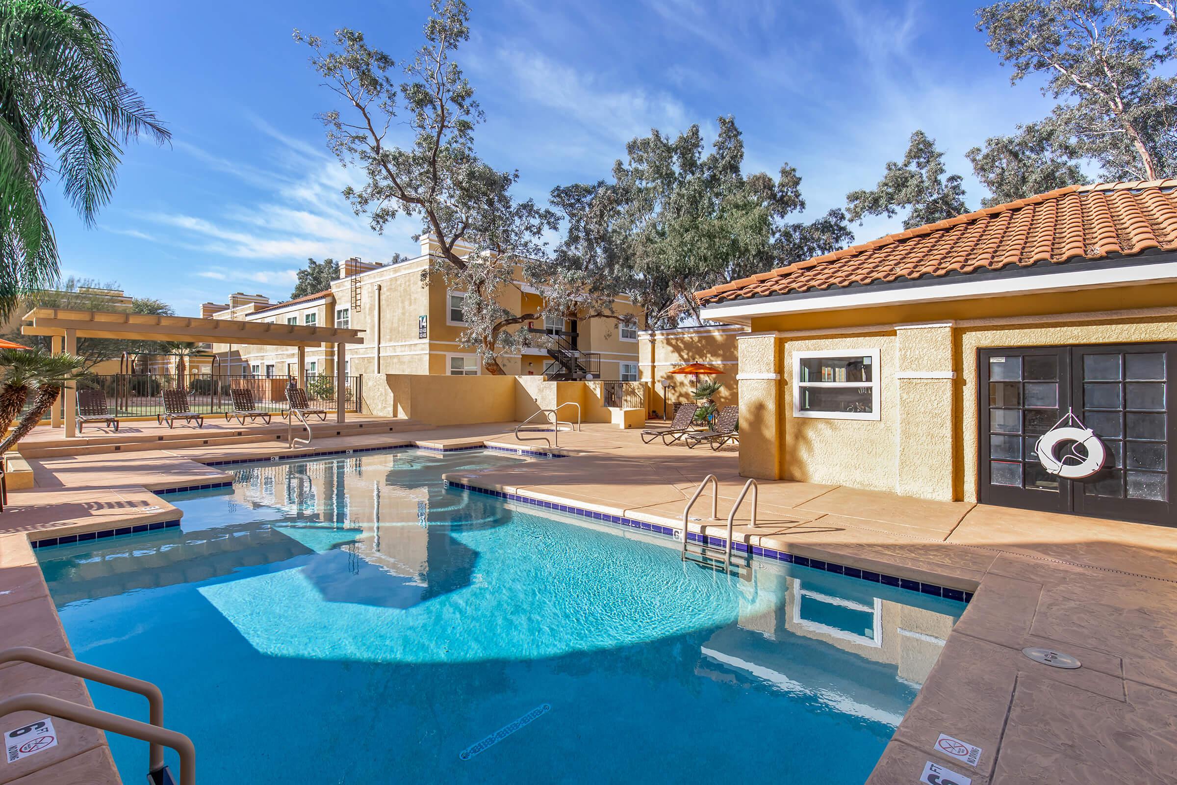 A serene pool area surrounded by palm trees, featuring a clear blue pool, lounge chairs, and a nearby building with a tiled roof. The scene is bright and sunny, showcasing a well-maintained outdoor space ideal for relaxation and leisure.