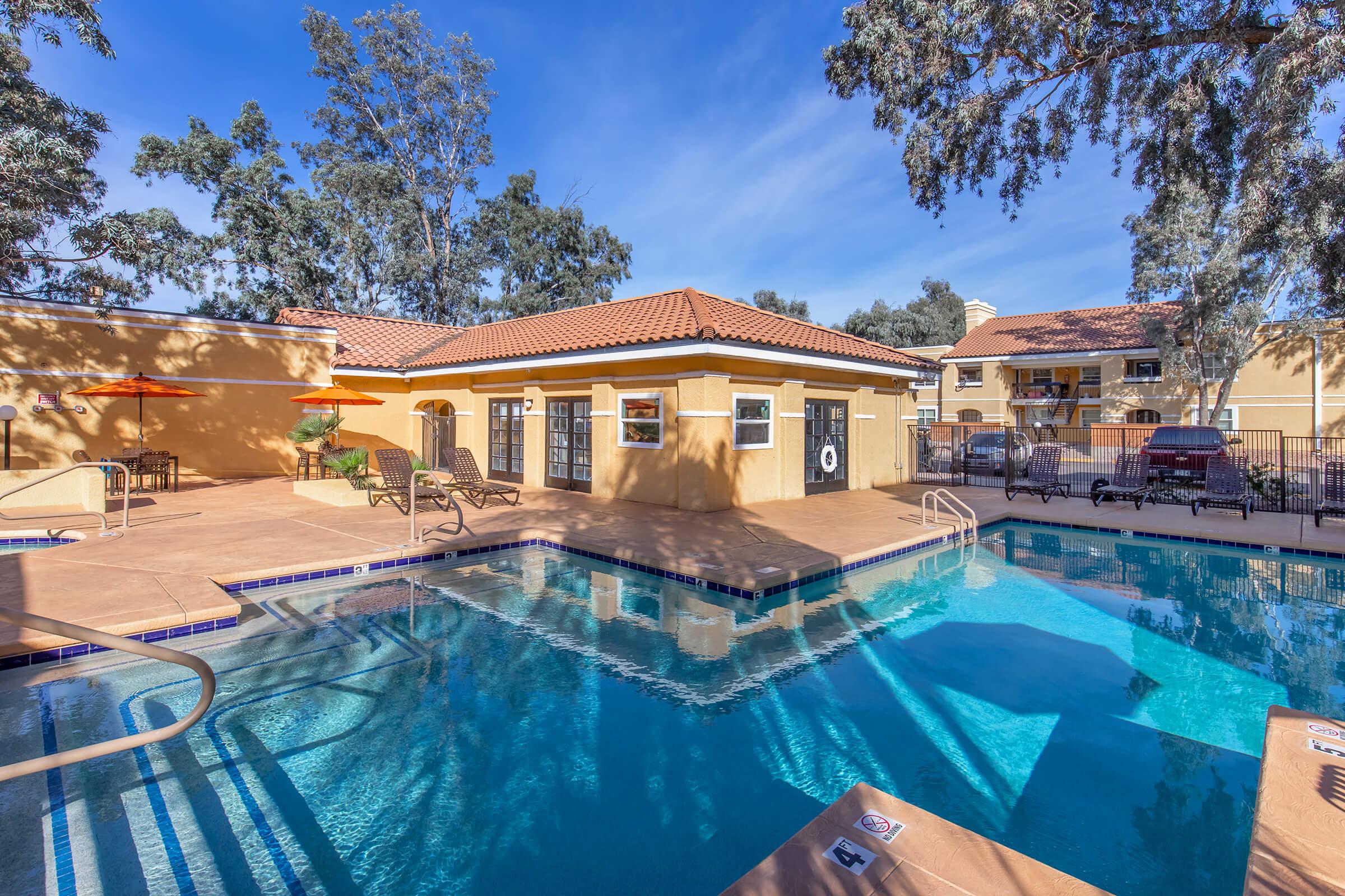 A sunny outdoor pool area featuring a clear blue water pool, lounge chairs, and red umbrellas. Surrounding the pool are well-maintained trees and a building with a warm beige exterior and a tiled roof. The scene suggests a relaxing atmosphere for leisure and enjoyment.