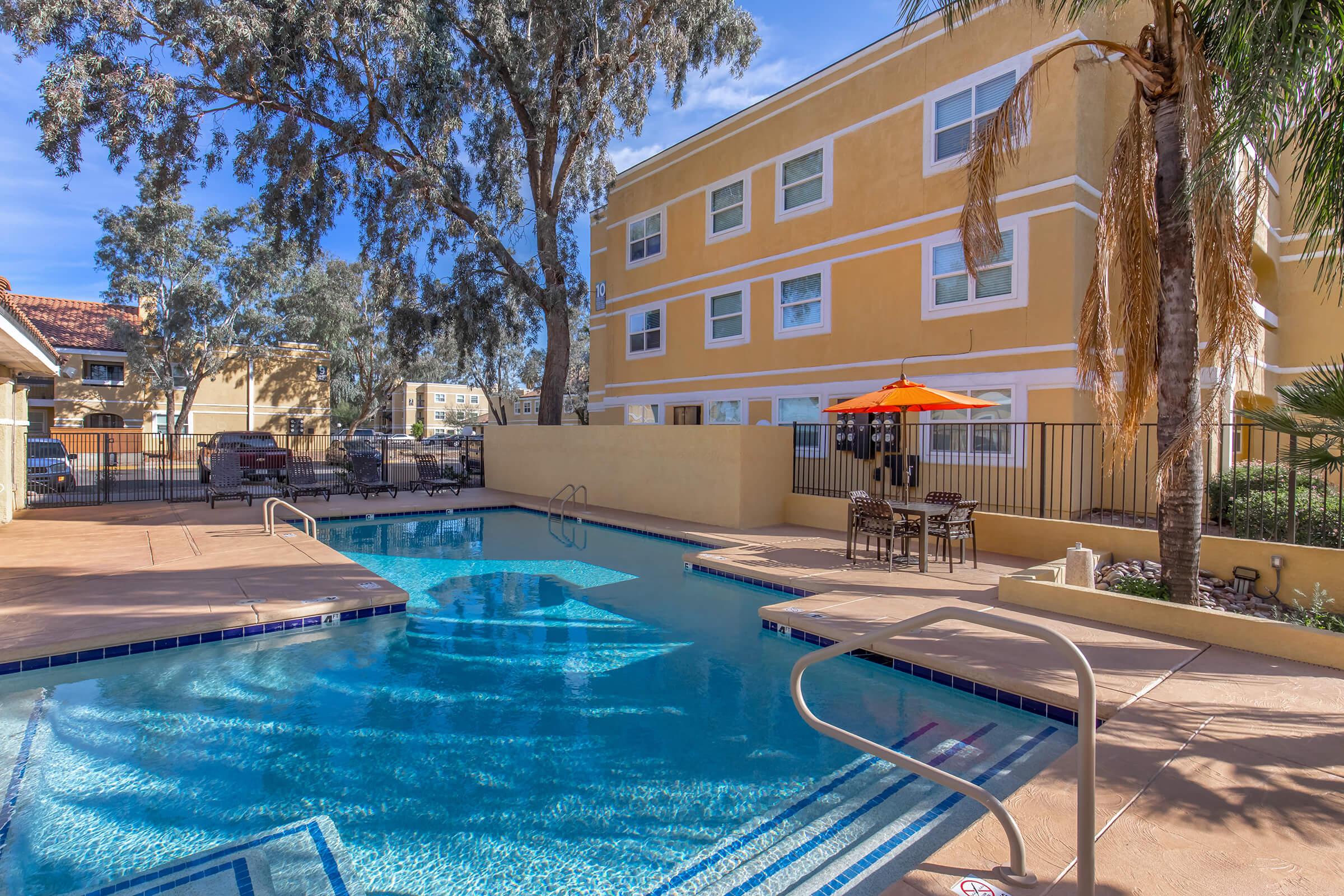A clean swimming pool surrounded by a patio area with lounge chairs and tables. In the background, a yellow multi-story building is visible, framed by trees. An orange umbrella adds a pop of color to the outdoor space. The scene captures a sunny and inviting atmosphere, perfect for relaxation.