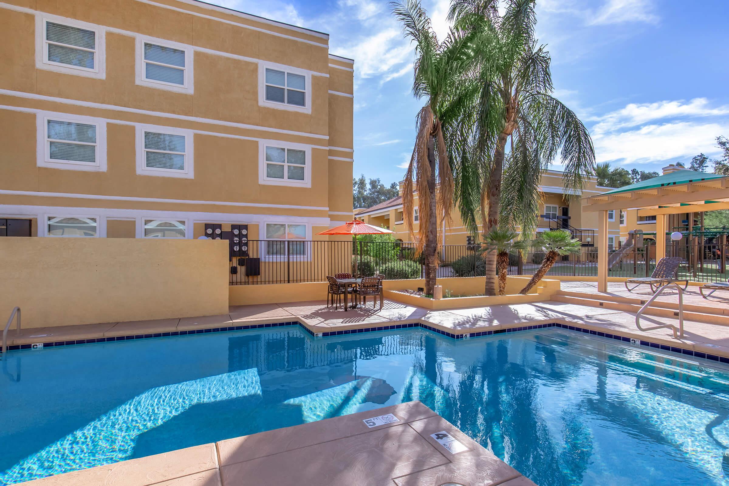 A sunny pool area with a clear blue swimming pool surrounded by lounge chairs and a shaded seating area with a small table. Palm trees add greenery to the setting, and an orange umbrella provides shade. Surrounding buildings are partially visible in the background under a bright blue sky.
