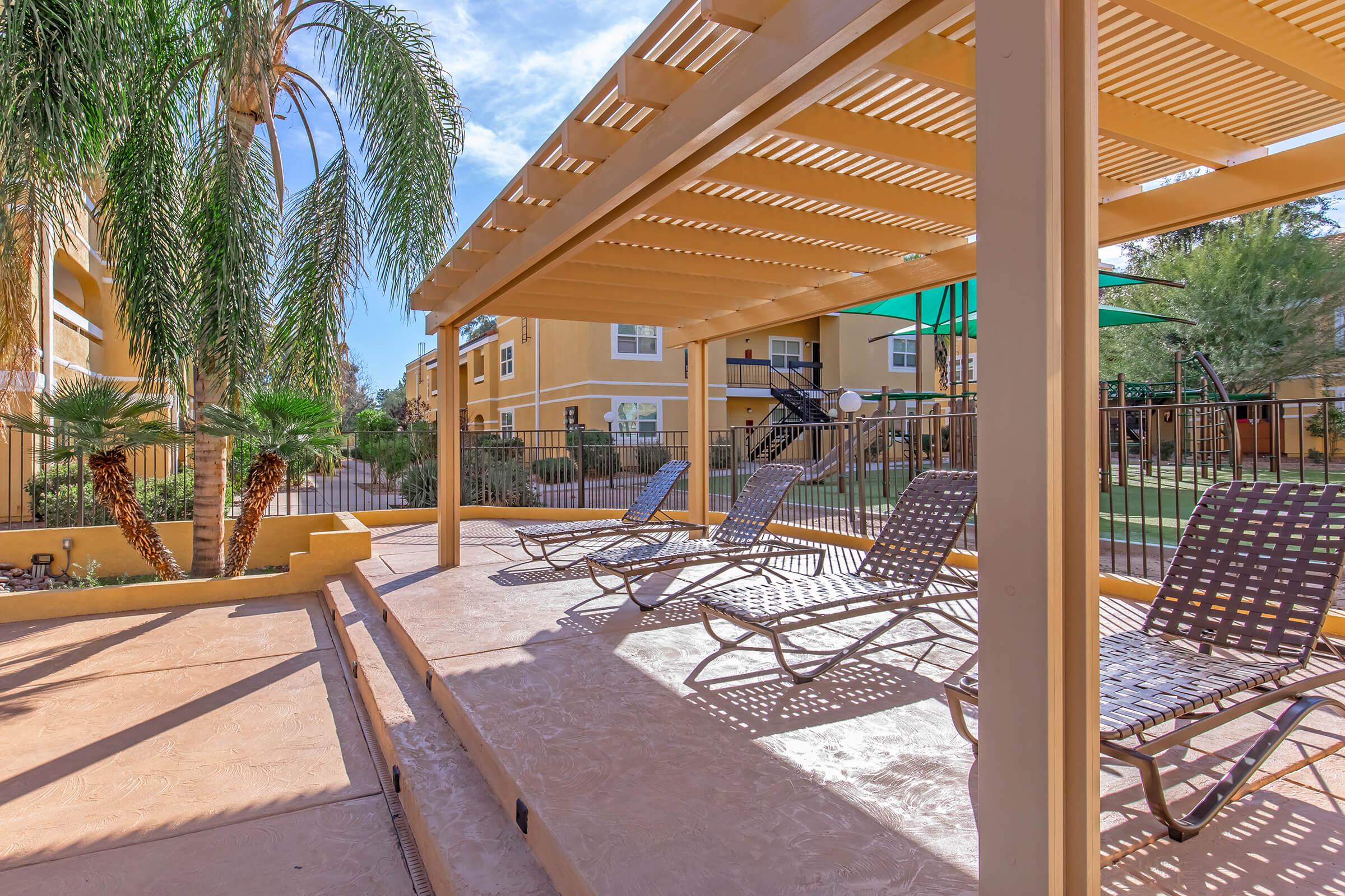 Lounging area with several reclining chairs under a large pergola, surrounded by tropical plants. The background features multi-story beige apartment buildings and green umbrellas, with a well-maintained lawn in a sunny outdoor setting.