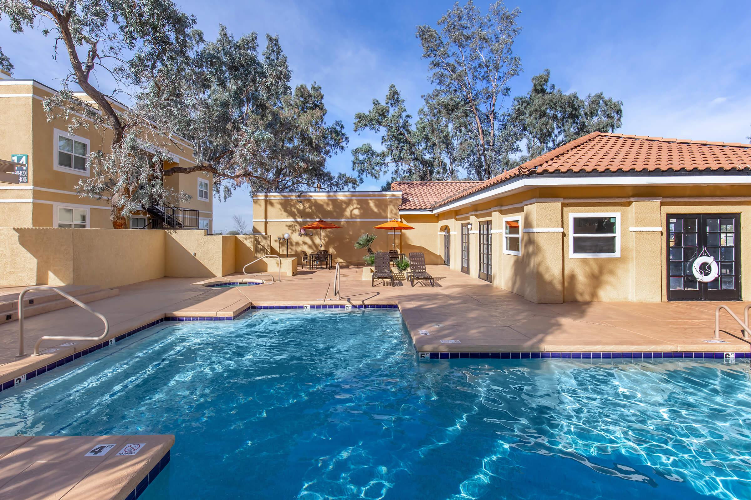 A bright outdoor pool area surrounded by trees, featuring a clear blue swimming pool, lounge chairs, and orange patio umbrellas. Nearby, there is a poolside building with large windows and a tiled roof, providing a relaxing atmosphere for residents and guests.