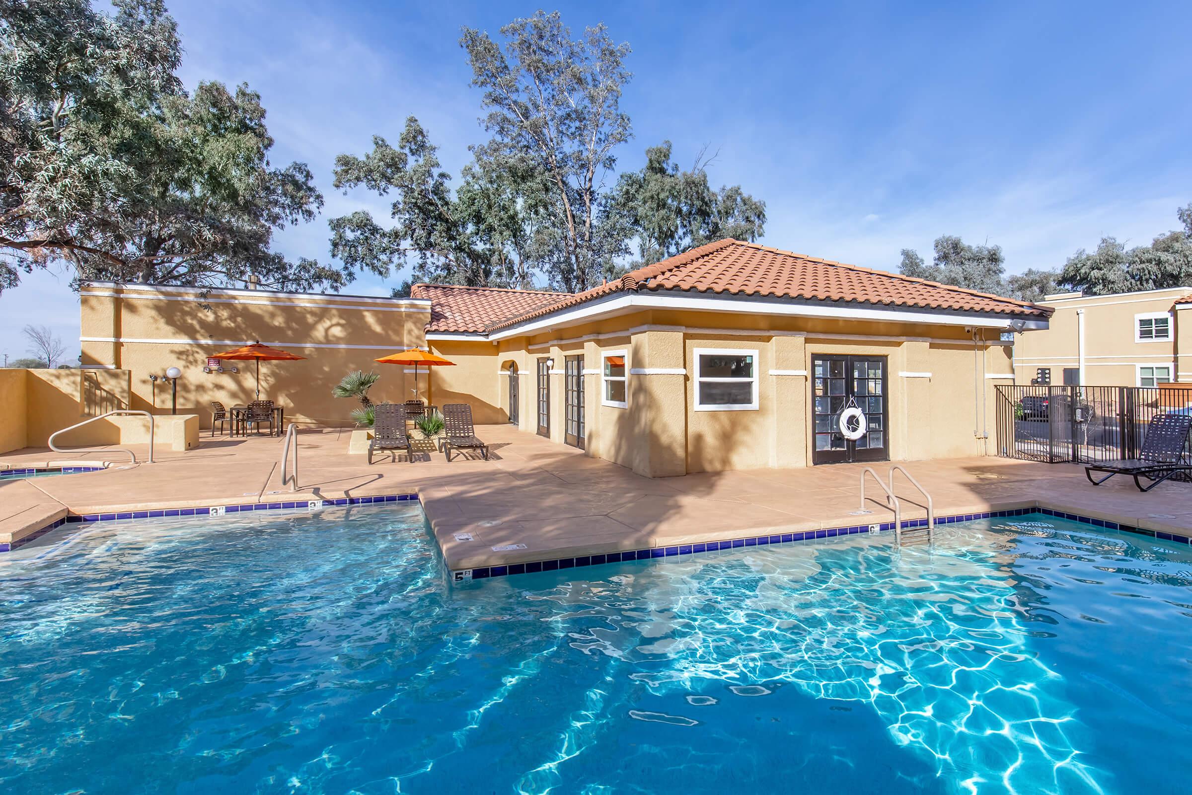 A swimming pool surrounded by a patio area with lounge chairs and umbrellas. There is a building with a tan exterior and a red-tiled roof adjacent to the pool. Lush trees provide shade in the background, and the sky is clear with a few clouds.