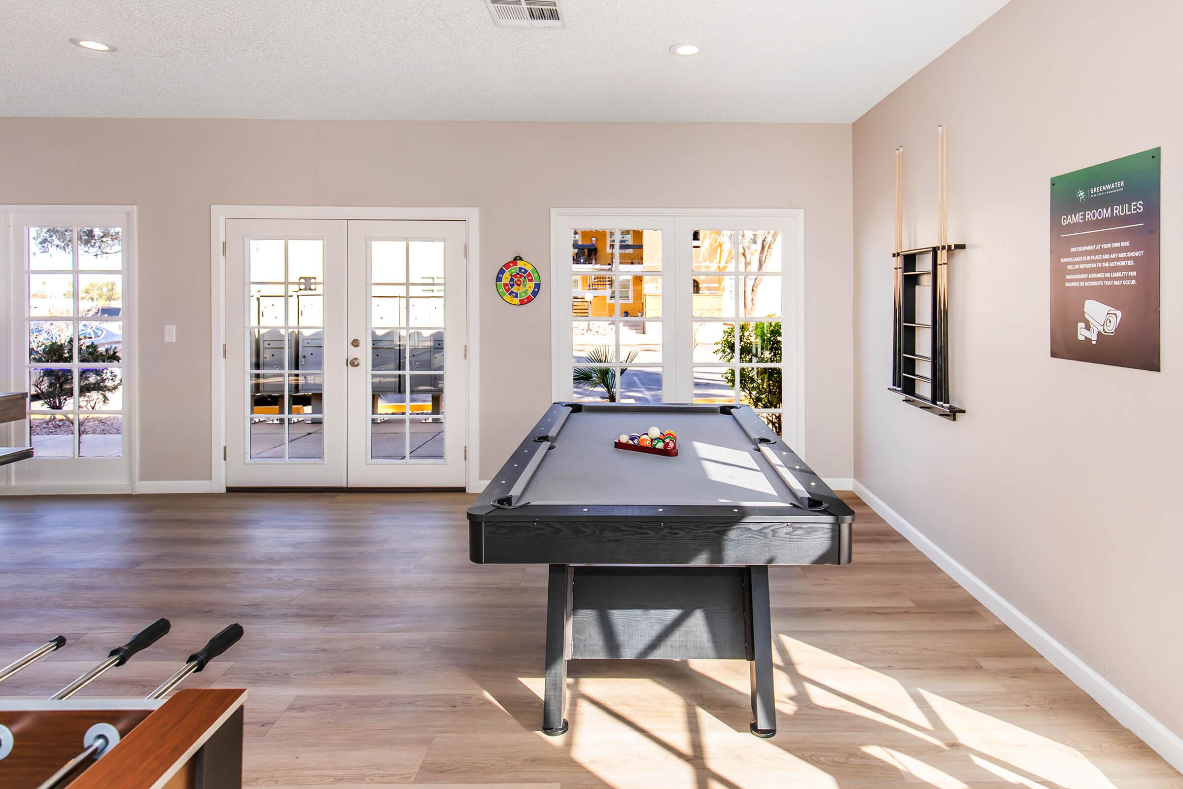 A bright, spacious game room featuring a black pool table with balls on it, large windows letting in natural light, and a foosball table in the foreground. A sign with game rules is mounted on the wall, and there’s colorful decor on the door. The floor is light wood, creating a welcoming atmosphere.