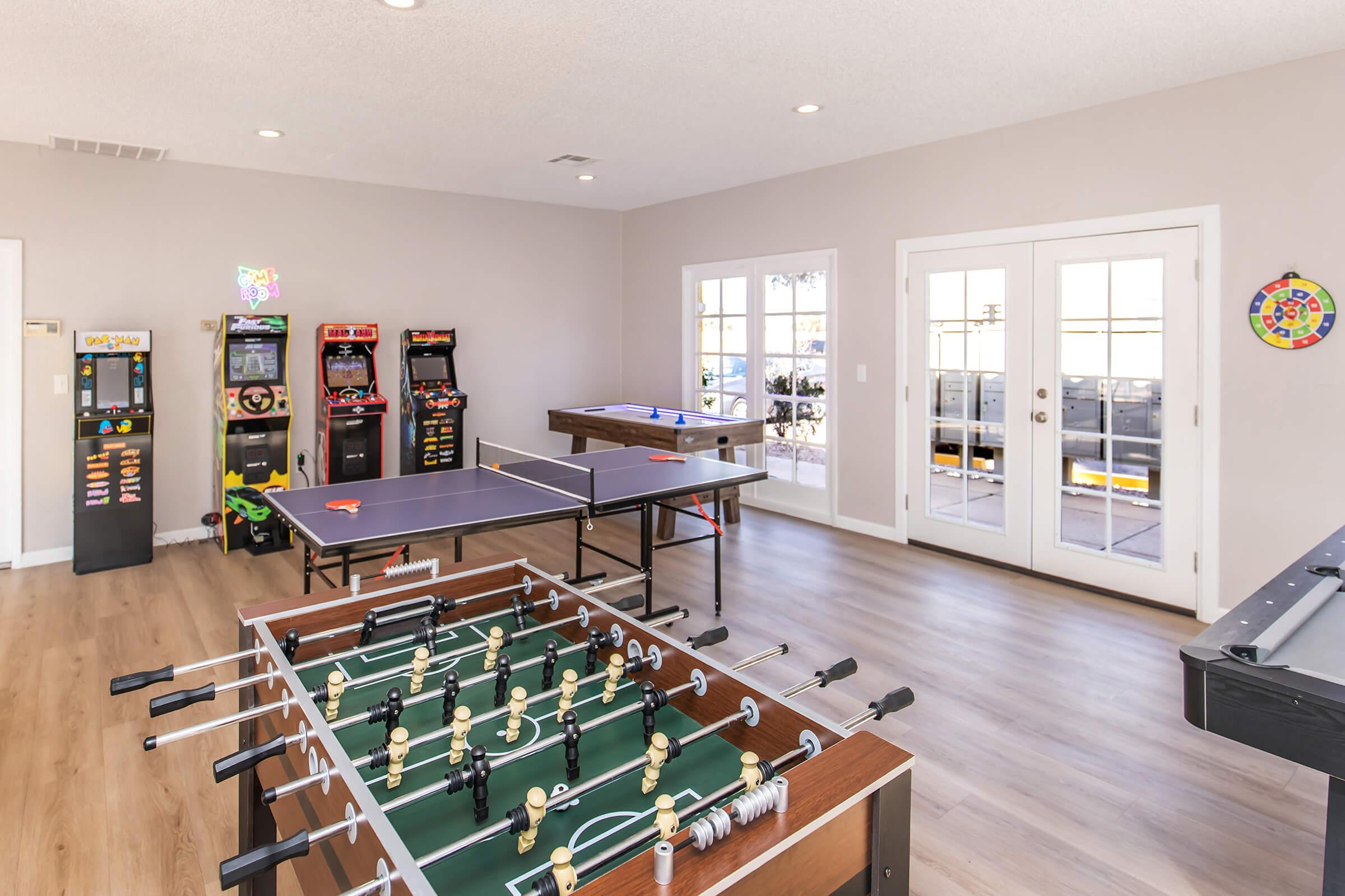 A spacious game room featuring a foosball table, ping pong table, and several arcade game machines. Large windows allow natural light, and there are double doors leading outside. The room has a modern design with wooden flooring and a cheerful atmosphere, perfect for recreational activities.