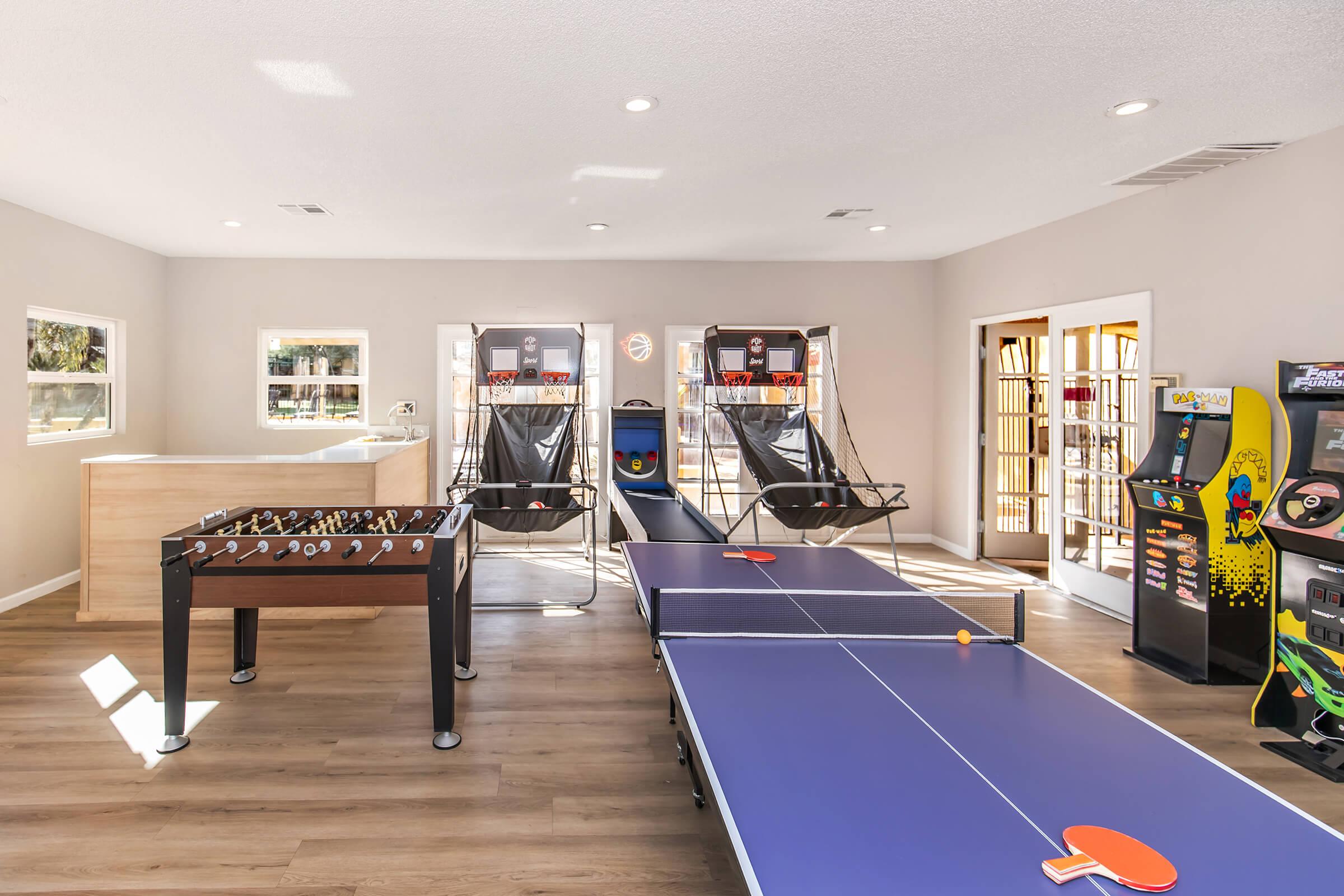 A well-lit game room featuring a pool table, arcade machines, and a large screen. The room has light-colored walls and a modern design, with a vibrant carpet and comfortable seating, creating an inviting atmosphere for gaming and entertainment. Large windows provide natural light.