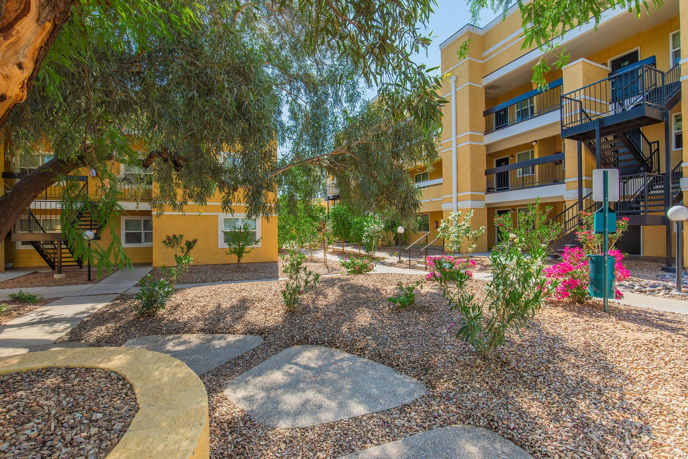 A well-maintained courtyard featuring landscaped greenery, pathways, and flowering plants. Surrounding the area are two-story apartment buildings in a warm yellow hue, with balconies and staircases visible. Sunlight filters through trees, creating a tranquil outdoor space.