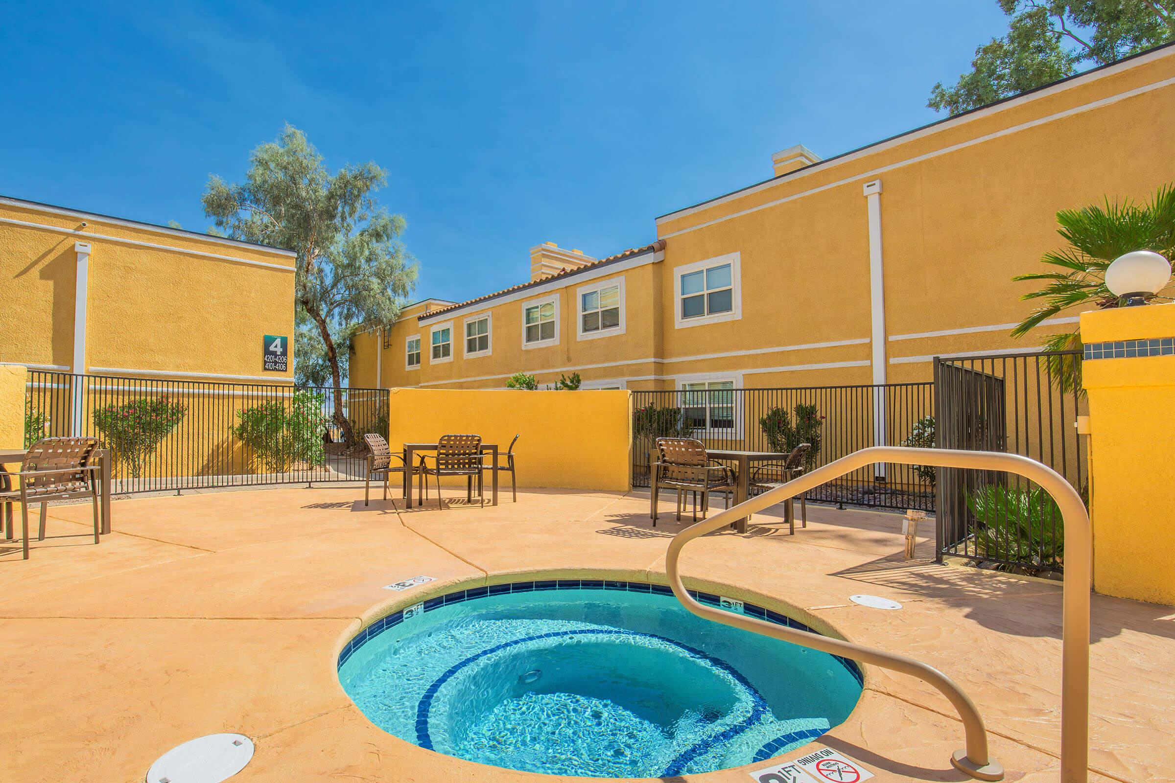 A hot tub surrounded by outdoor seating and palm trees, with a sunny blue sky and yellow buildings in the background. The area is fenced and features a clean, inviting ambiance, ideal for relaxation.