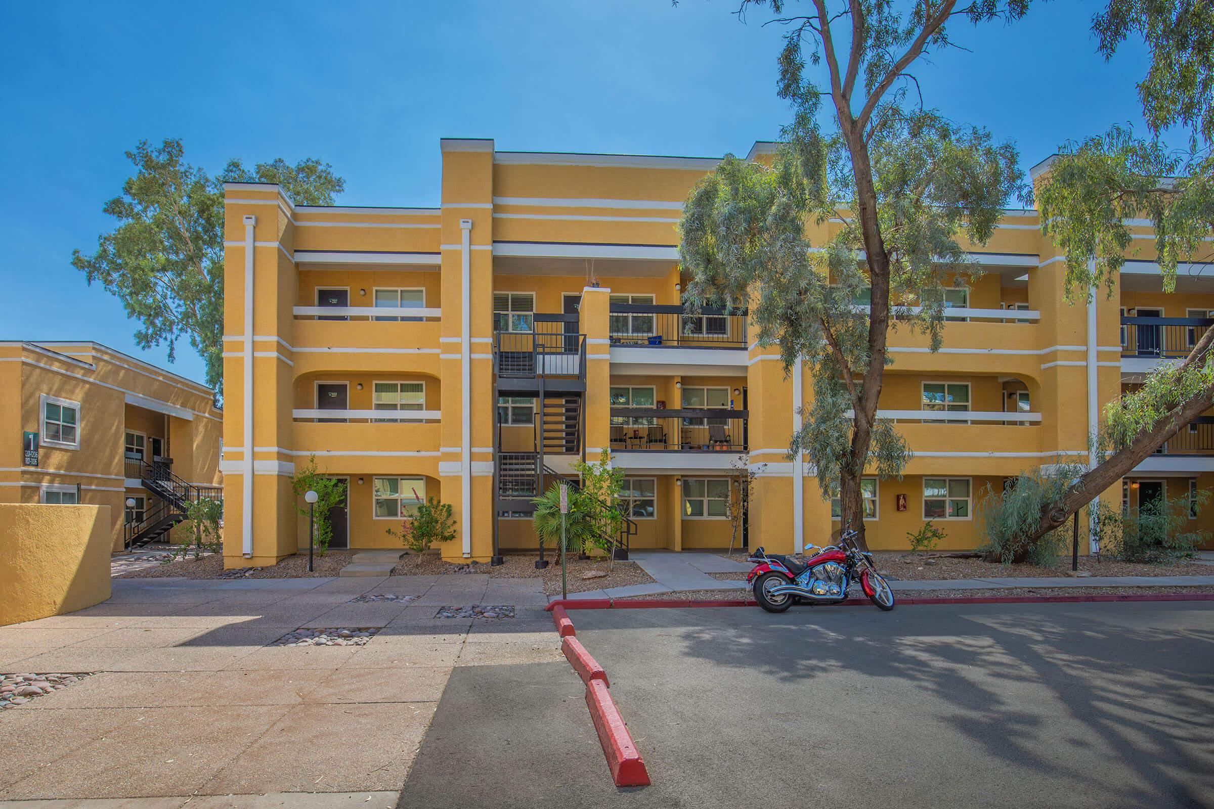 A three-story yellow apartment building featuring balconies, surrounded by trees and landscaping. In the foreground, a red motorcycle is parked along a paved path, with clear blue skies above.