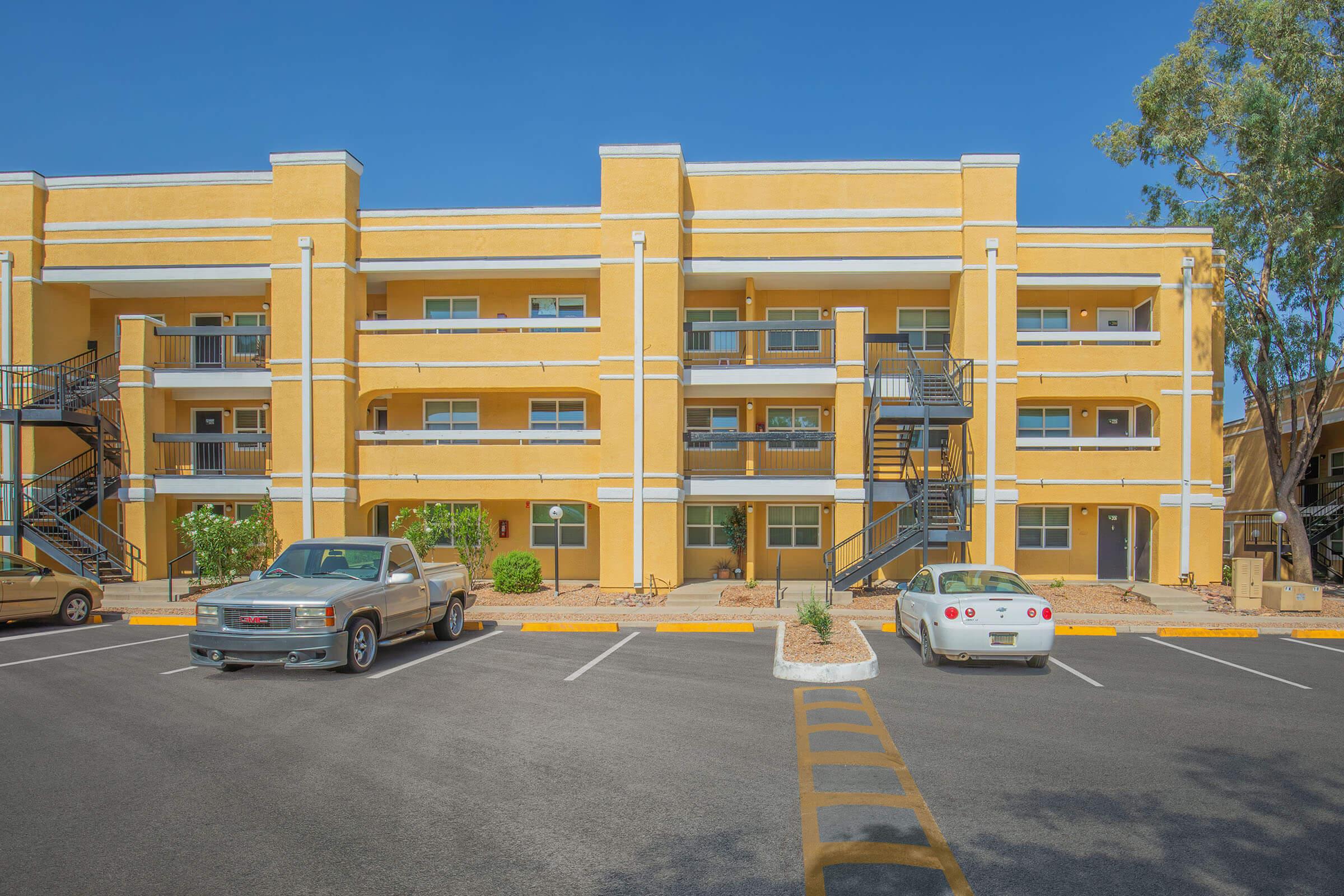 A large, yellow residential building with multiple levels and balconies. The structure features two vehicles parked in front: a gray pickup truck and a white sports car. Surrounding the building are landscaped areas with shrubs and small trees under a clear blue sky.