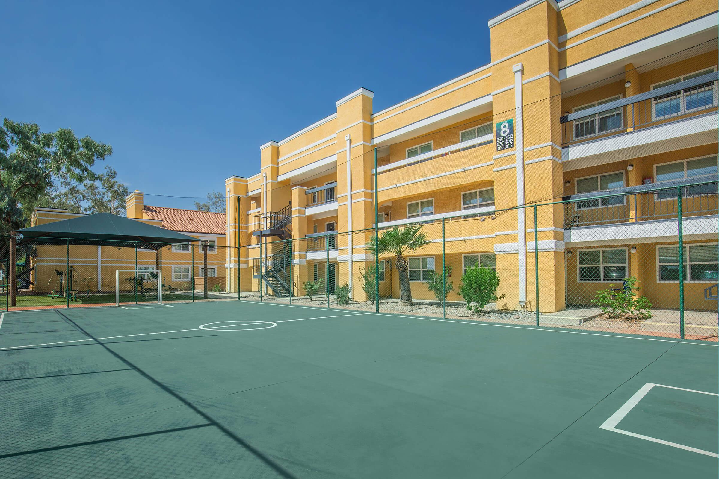 View of a green basketball court in an apartment complex. The court is enclosed by a fence, with yellow apartment buildings in the background. There are trees and shrubs nearby, and a shaded seating area to the side. The sky is clear and blue, creating a bright atmosphere.