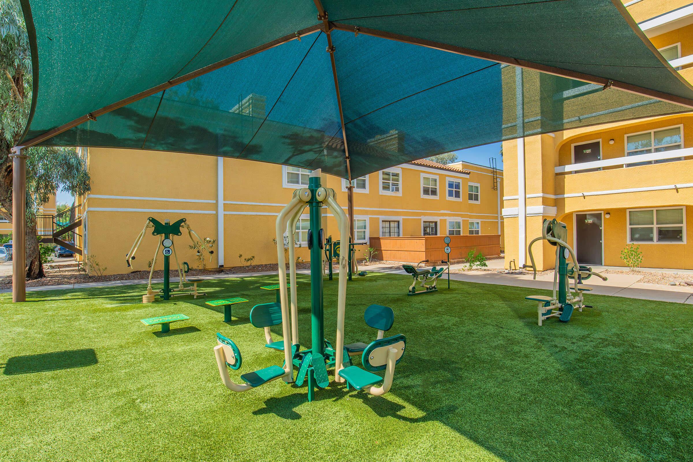 A well-maintained outdoor fitness area shaded by a large canopy. The space features various exercise equipment such as pull-up bars and seated leg press machines, situated on artificial grass. Surrounding the area are yellow apartment buildings with large windows.