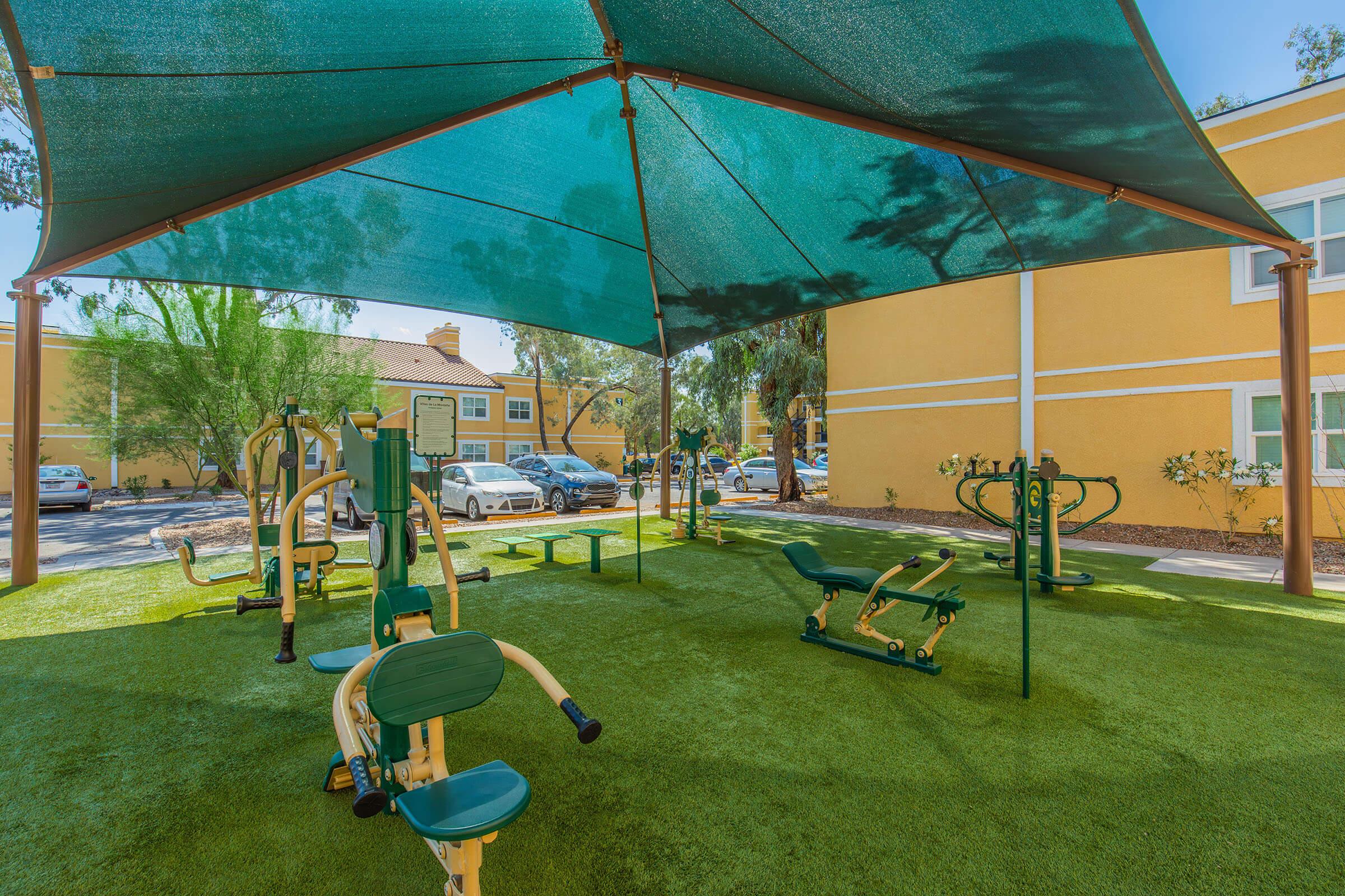 Outdoor fitness area featuring various exercise equipment under a large shaded canopy. The space is surrounded by greenery, with residential buildings in the background. The ground is covered in artificial turf, providing a comfortable surface for workouts.