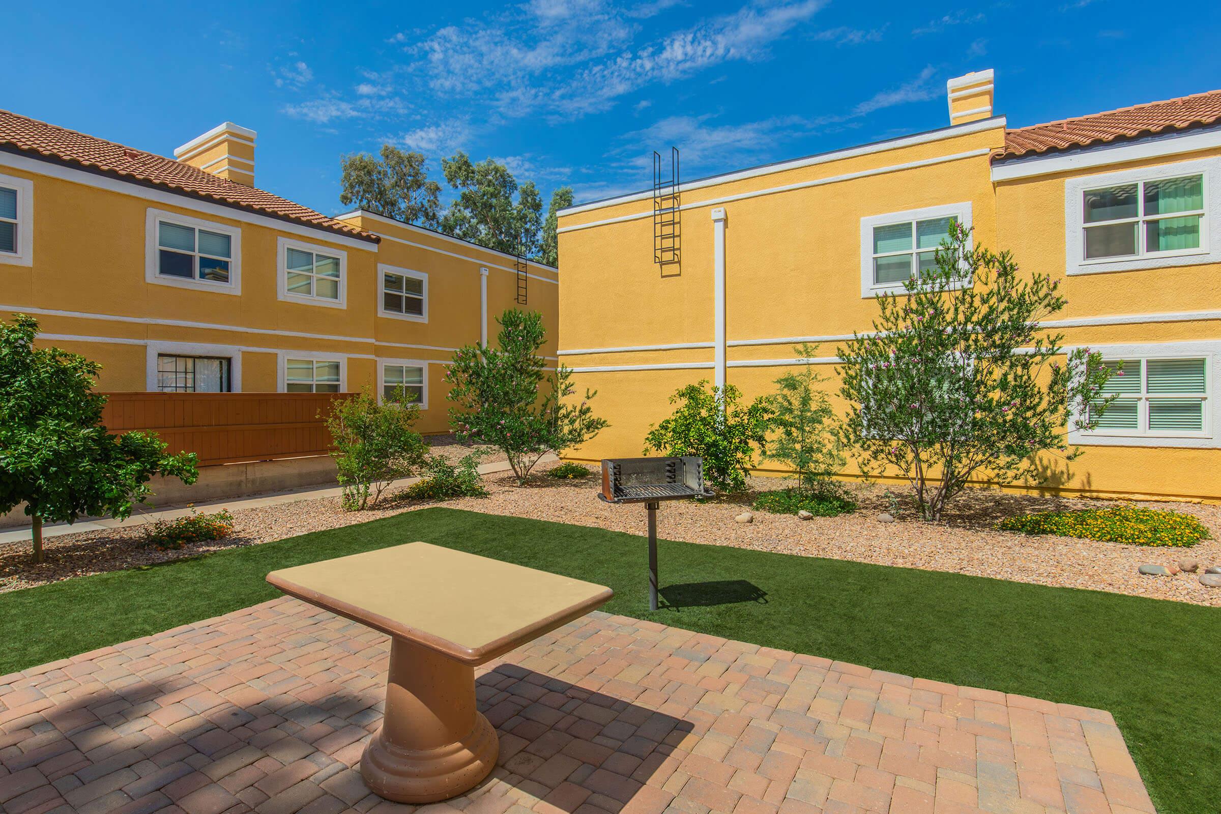 A sunny courtyard featuring a light yellow residential building, green shrubs, and a small grill area with a stone table. The ground is covered with pavers and artificial grass, surrounded by trees and plantings, under a clear blue sky.
