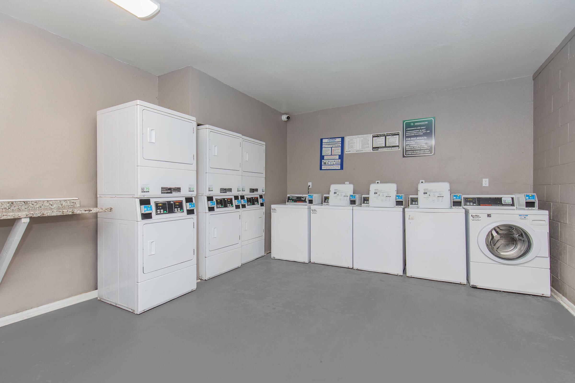 A clean, well-lit laundry room featuring multiple white washing machines and dryers. There is a countertop for folding clothes and informational signage on the wall. The floor is gray, and the overall space is organized and functional.