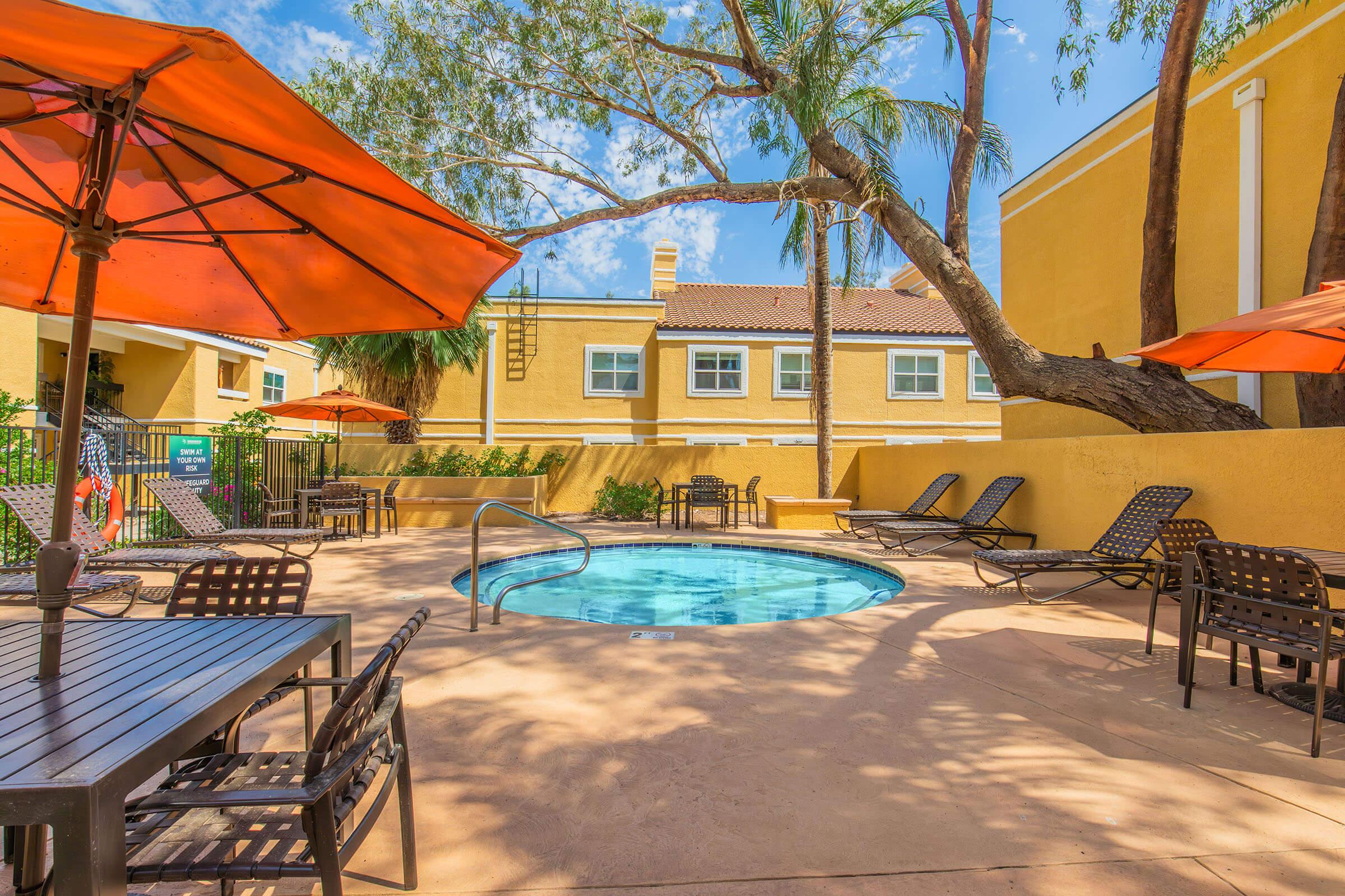 A pool area with a circular hot tub surrounded by lounge chairs and tables. Orange umbrellas provide shade, and palm trees enhance the tropical atmosphere. In the background, a yellow building with windows is visible under a clear blue sky.