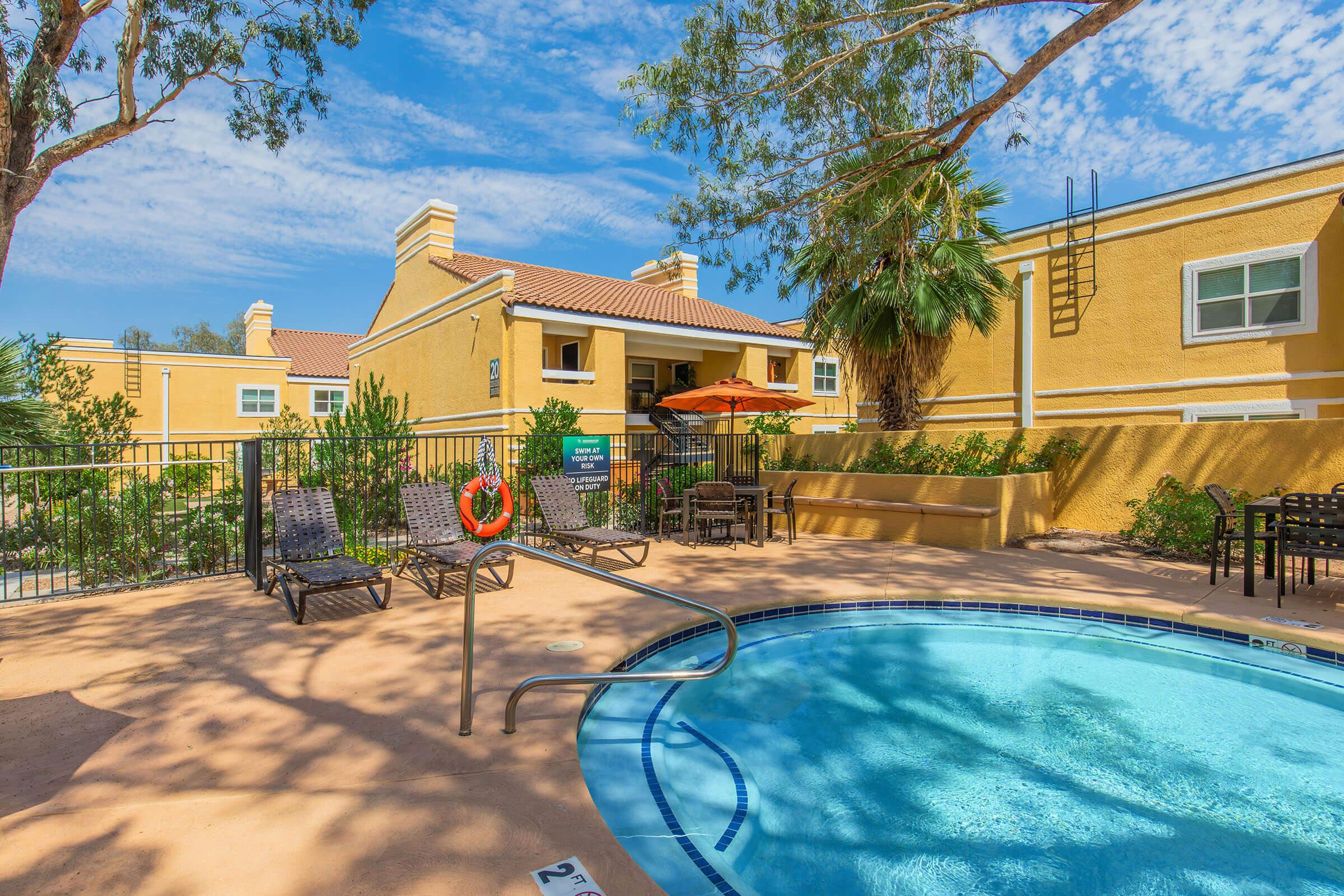 A sunny outdoor pool area with lounge chairs and an umbrella. Surrounding the pool are green plants, a few trees, and a building in the background. The scene is bright, showcasing a clear blue sky and inviting atmosphere. A life preserver is visible near the pool's edge.