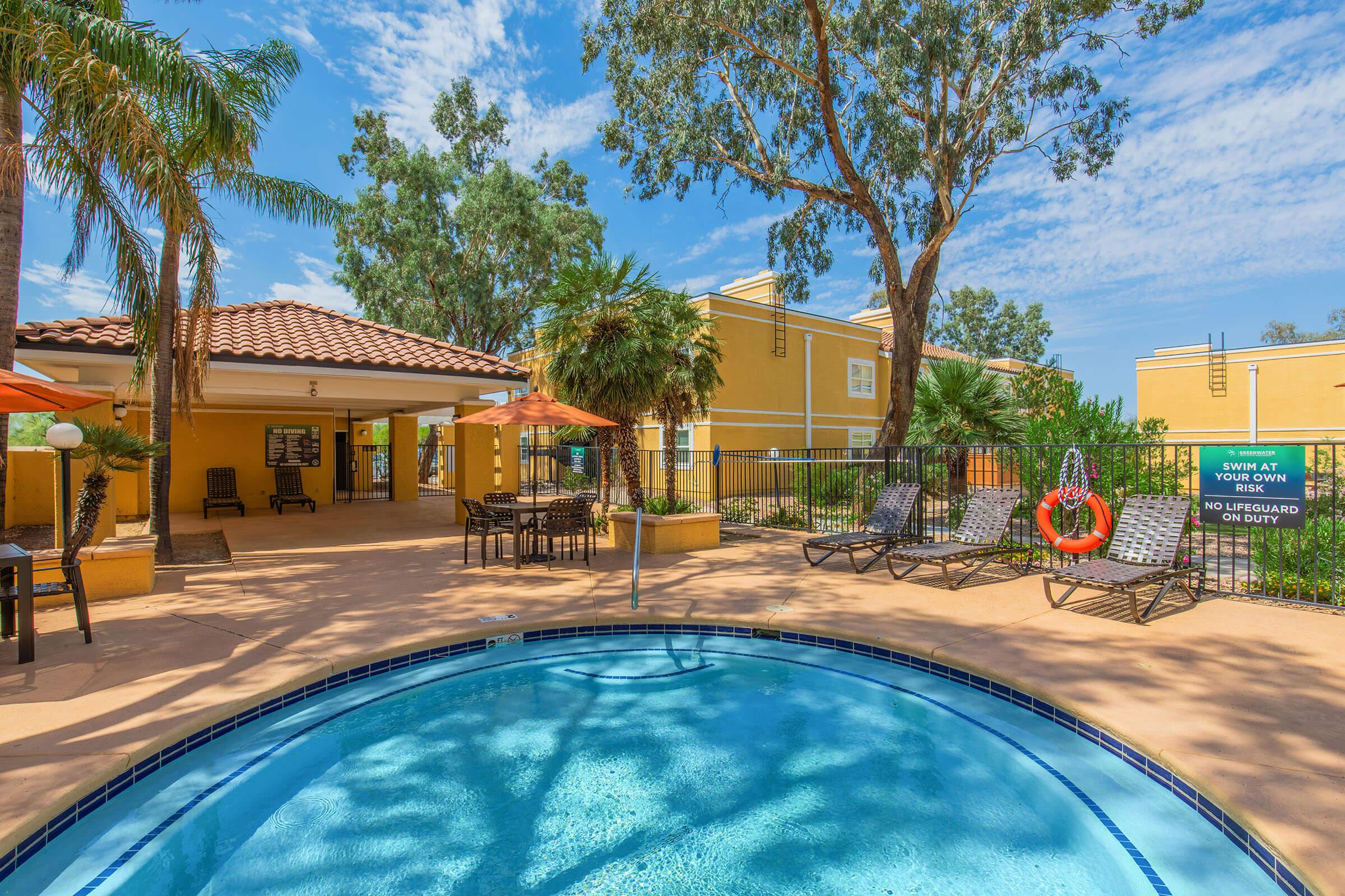 A sunny outdoor area featuring a swimming pool surrounded by lounge chairs and umbrellas. In the background, there are palm trees and a yellow building. A sign indicates pool safety rules. The scene conveys a relaxing atmosphere, ideal for leisure and enjoyment.