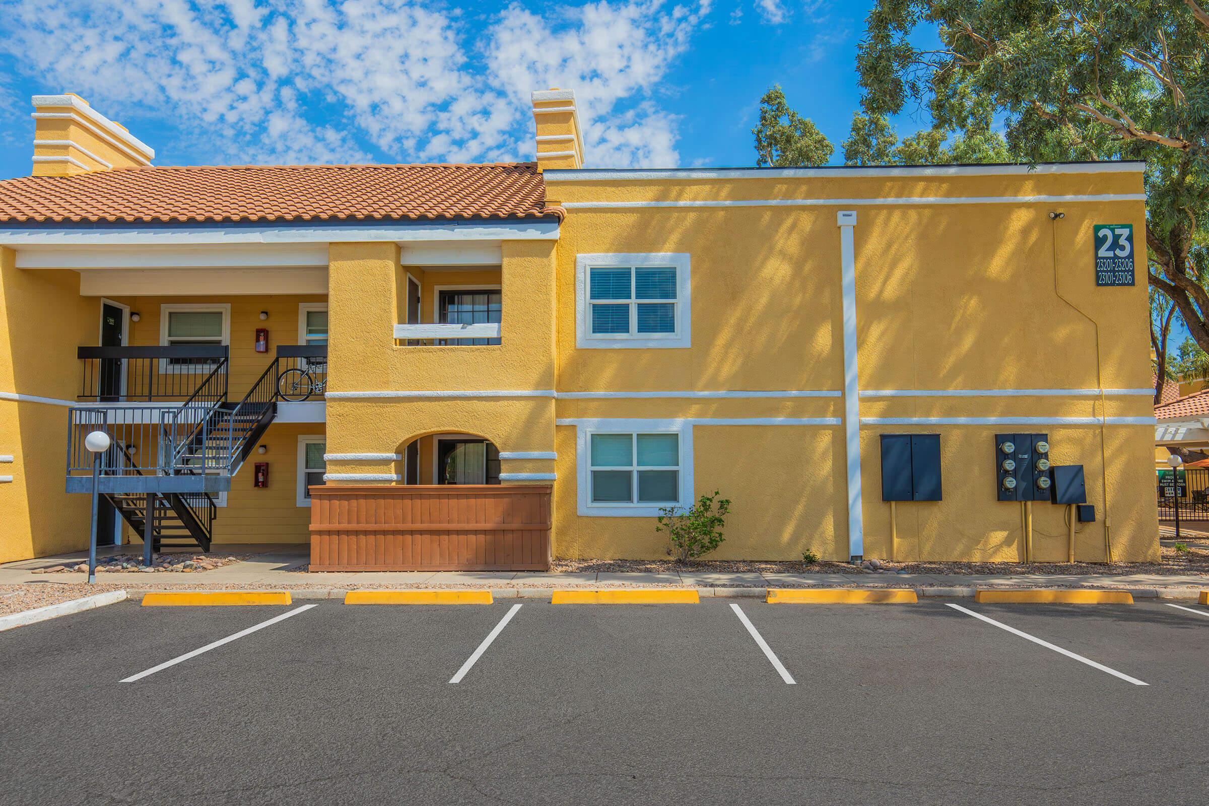 A two-story yellow building with a tiled roof and white trim. It features a staircase leading to the upper level, numbered 23 on the side. The foreground has a parking area with several painted spaces, and there are green trees nearby under a partly cloudy blue sky.