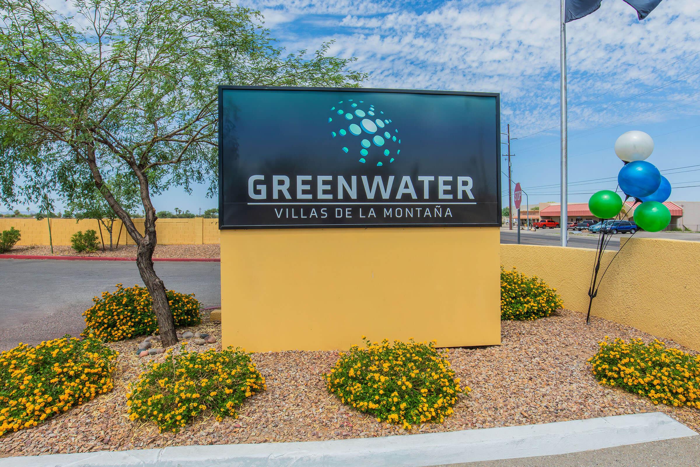 Sign for "Greenwater Villas de la Montaña" displaying the name prominently, surrounded by colorful flower beds and decorative balloons. The background features a clear blue sky and some distant buildings, indicating a welcoming residential area.