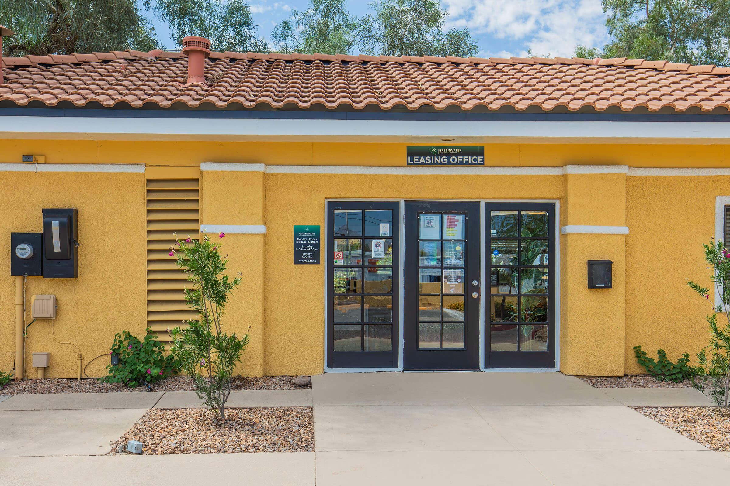 Exterior view of a leasing office with a bright yellow facade, featuring double glass doors, windows, and a tiled roof. There are plants and landscaping near the entrance, as well as a mailbox and signage displayed on the building. The setting appears well-maintained and inviting.