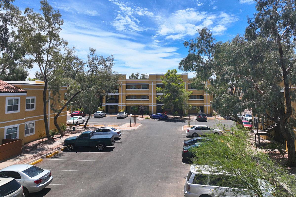A view of an apartment complex surrounded by trees, featuring two buildings with yellow exteriors. A parking lot is visible in the foreground with several cars, and bright blue skies with scattered clouds create a sunny atmosphere.