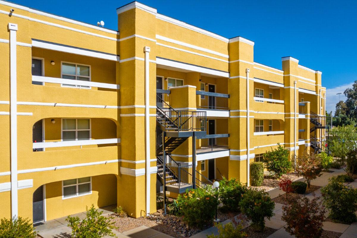 Bright yellow multi-story apartment building with a modern design. The exterior features multiple balconies, black metal staircases, and landscaped pathways with shrubs and trees visible in the foreground. Clear blue sky overhead creates a vibrant atmosphere.