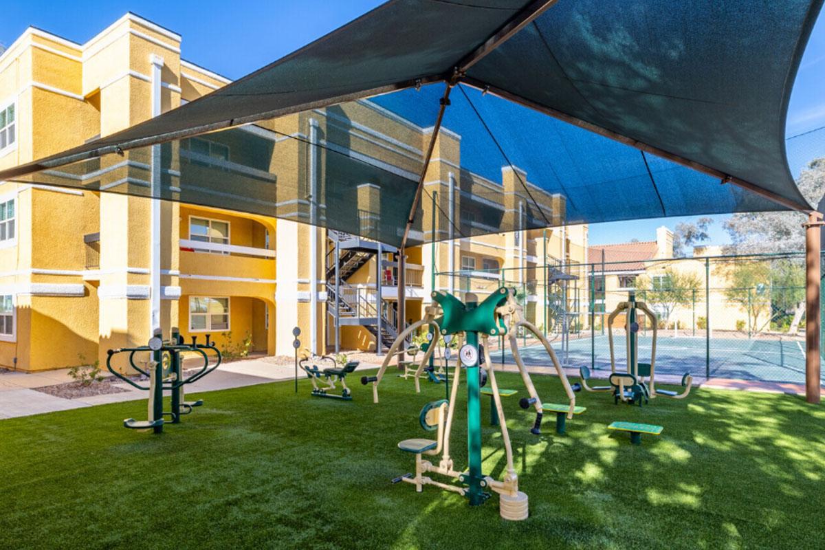 Outdoor fitness area featuring various exercise equipment under a shaded canopy. The setting includes green artificial turf and nearby residential buildings. The space is designed for community engagement and physical activity.