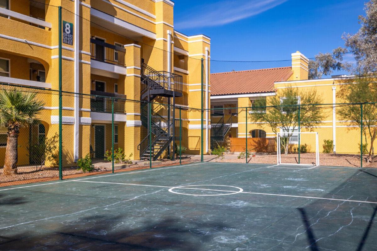 A view of a basketball court enclosed by a fence, located in an apartment complex with bright yellow buildings. The court shows worn lines and is surrounded by palm trees and other greenery under a clear blue sky.